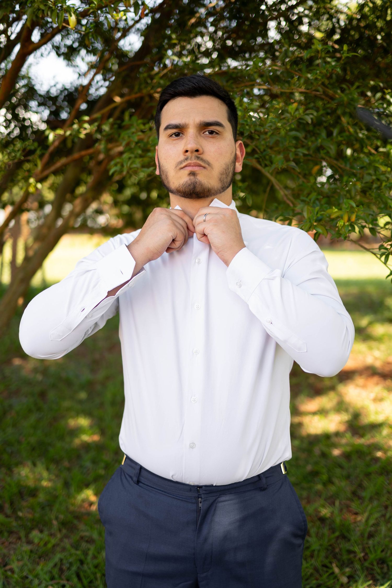 A young man in a white dress shirt adjusts his collar outdoors, standing near lush green trees.