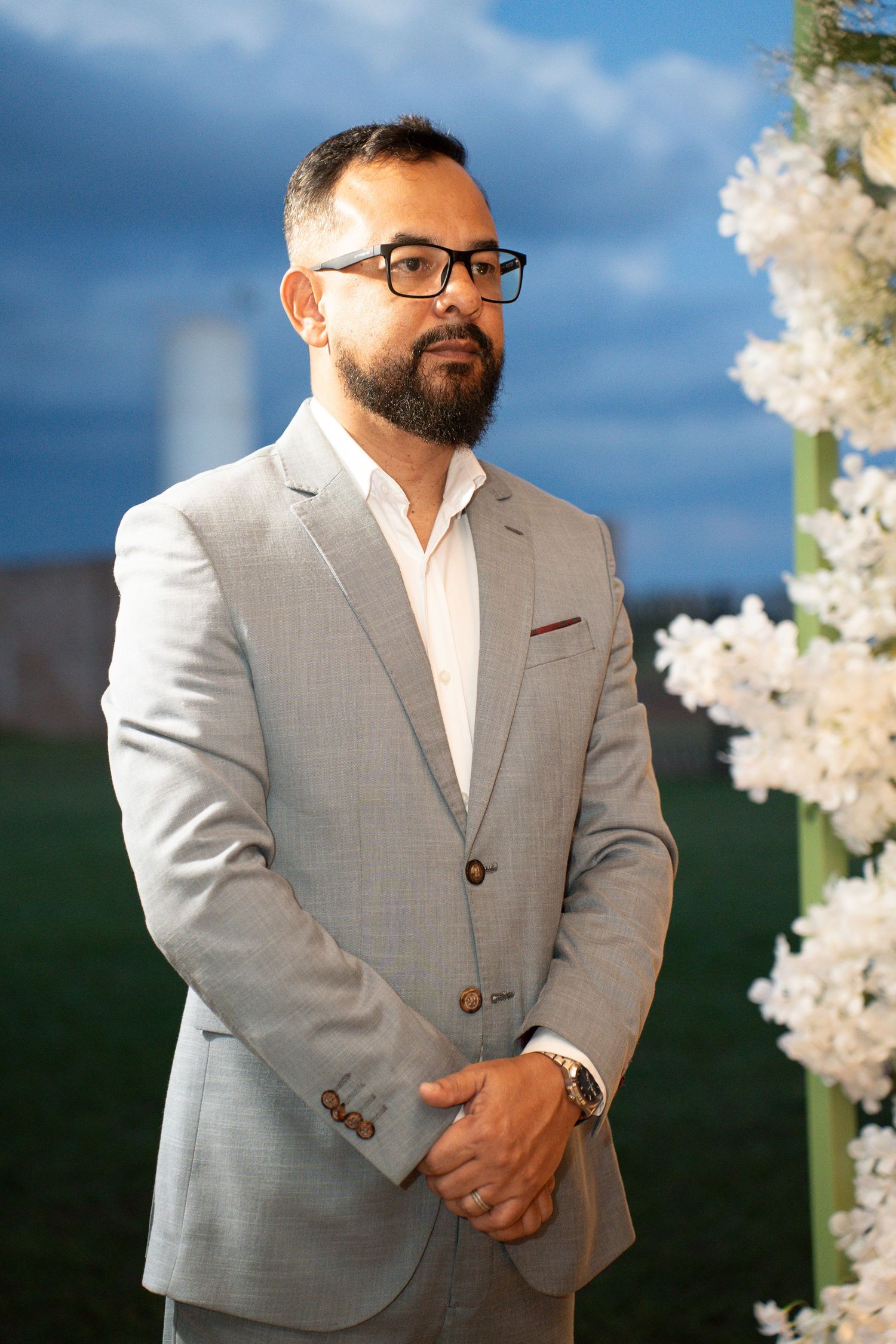A bearded man in glasses and a light gray suit stands outdoors near white flowers at a formal event.