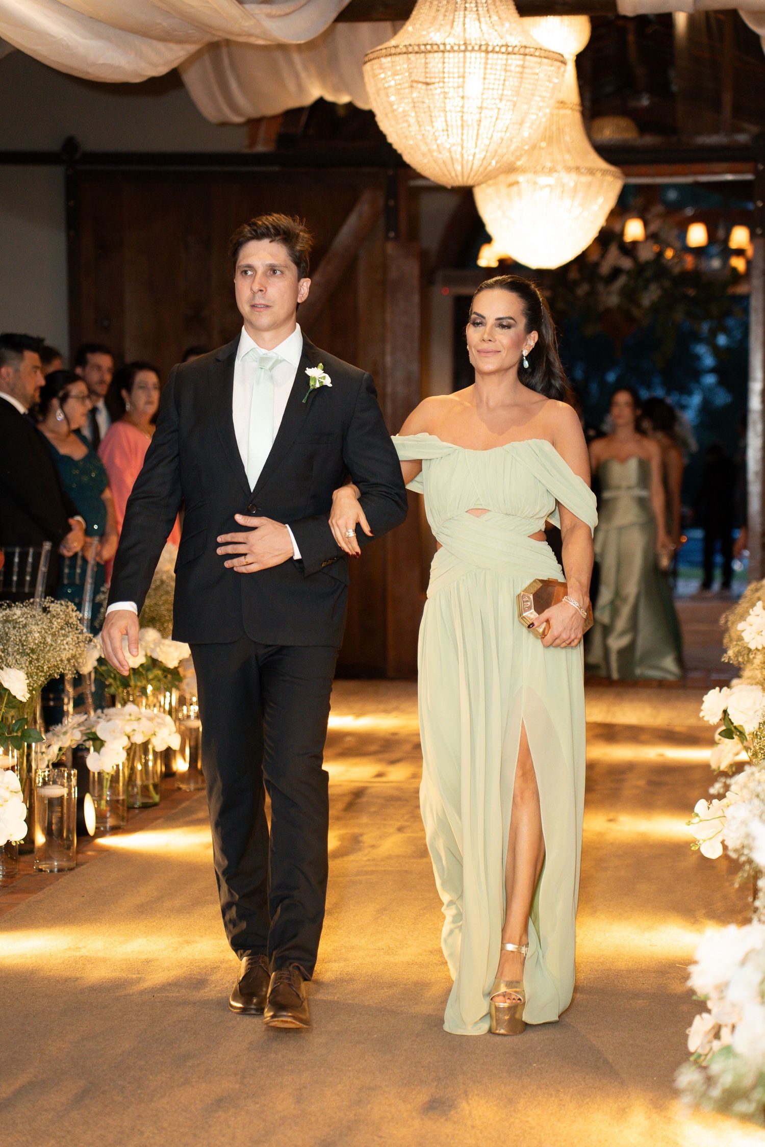 Elegant couple walking down wedding aisle, man in black tuxedo, woman in sage green off-shoulder gown with slit.
