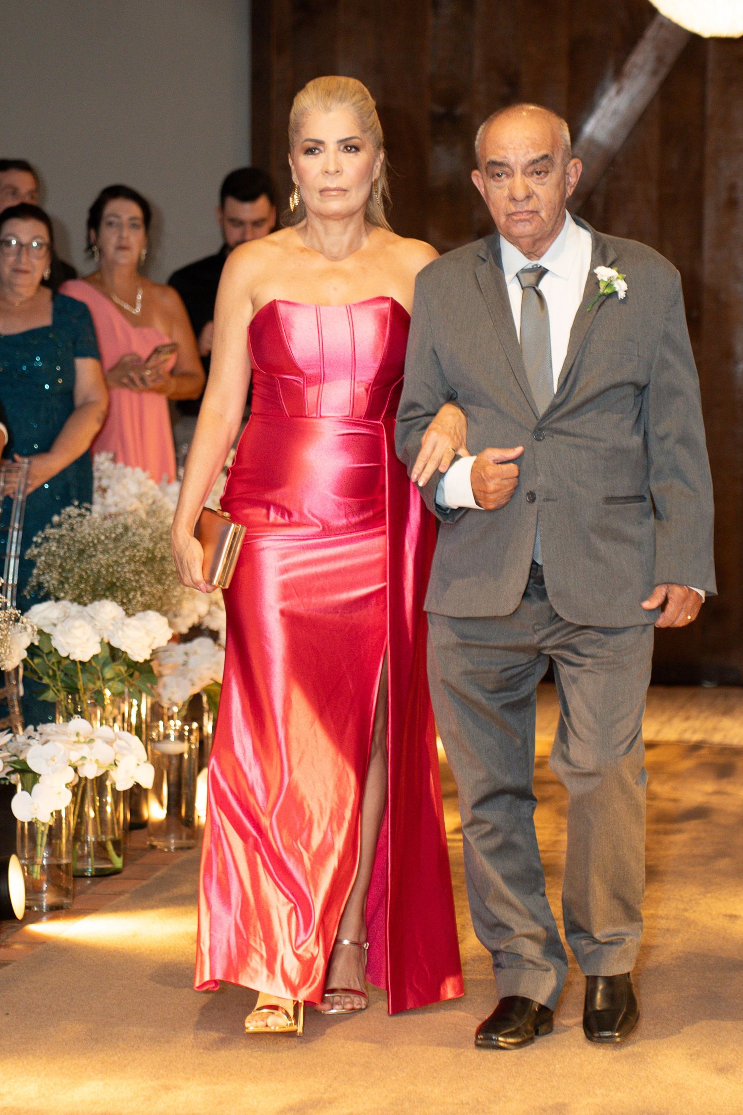 Elegant couple walking at a wedding, woman in red satin gown, man in gray suit with boutonniere.