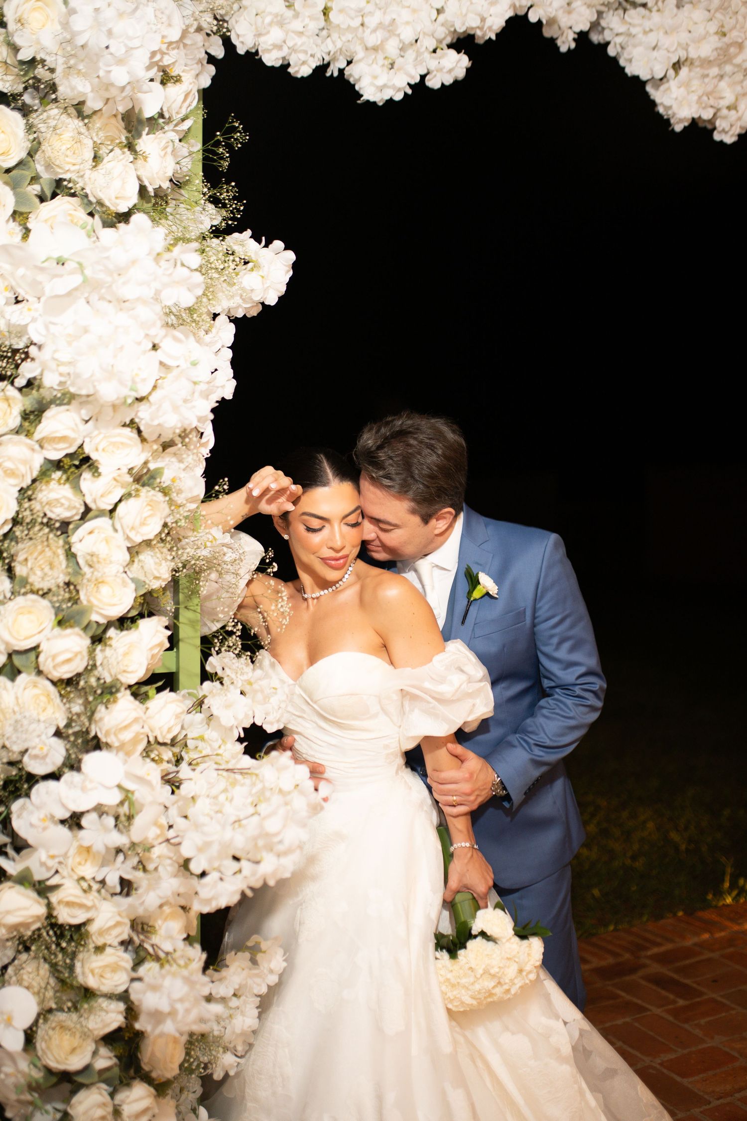 Bride and groom embrace under a stunning white floral arch at an elegant outdoor evening wedding ceremony.