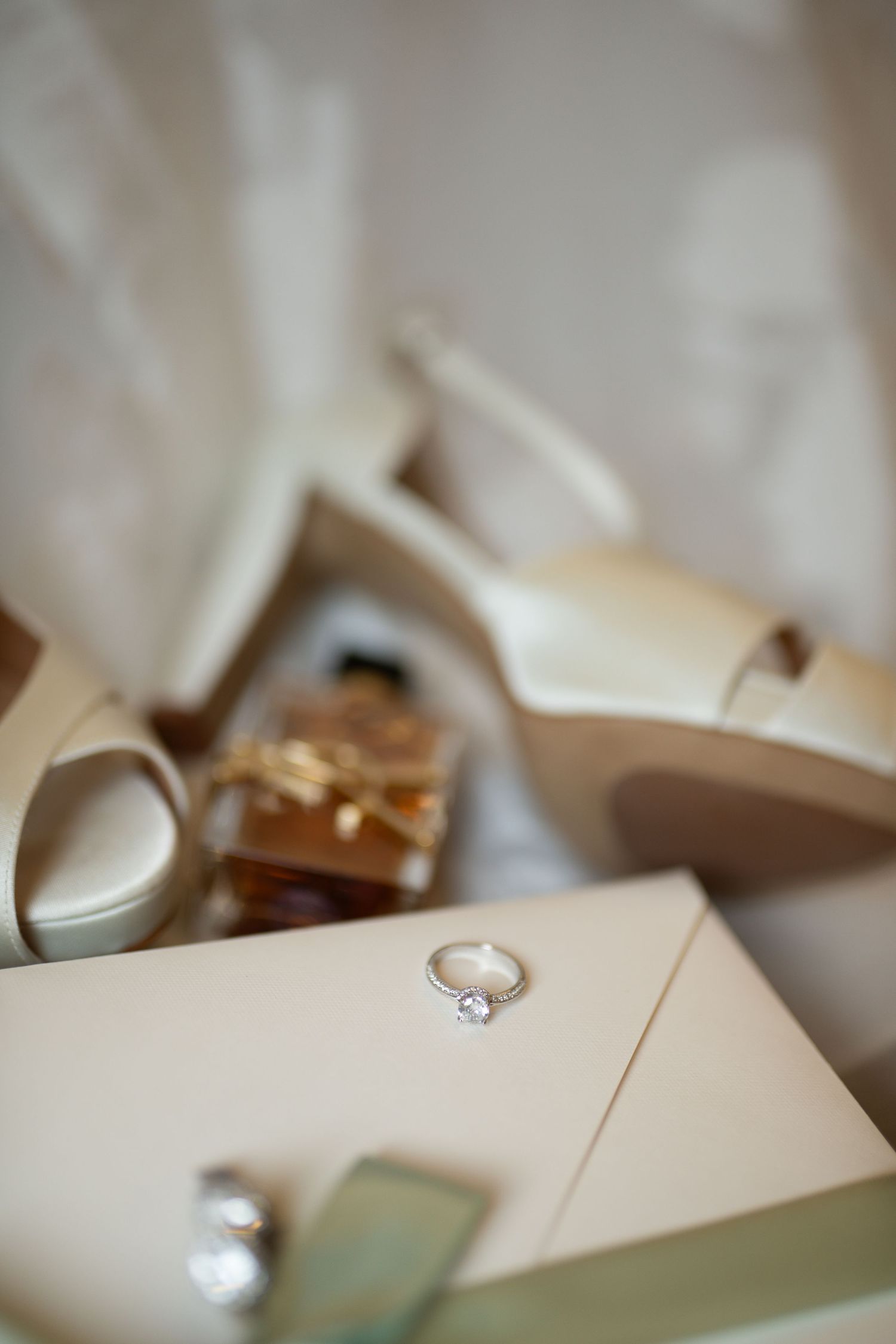 Elegant bridal flat lay featuring diamond engagement ring, cream heels, amber perfume bottle, and white envelope.