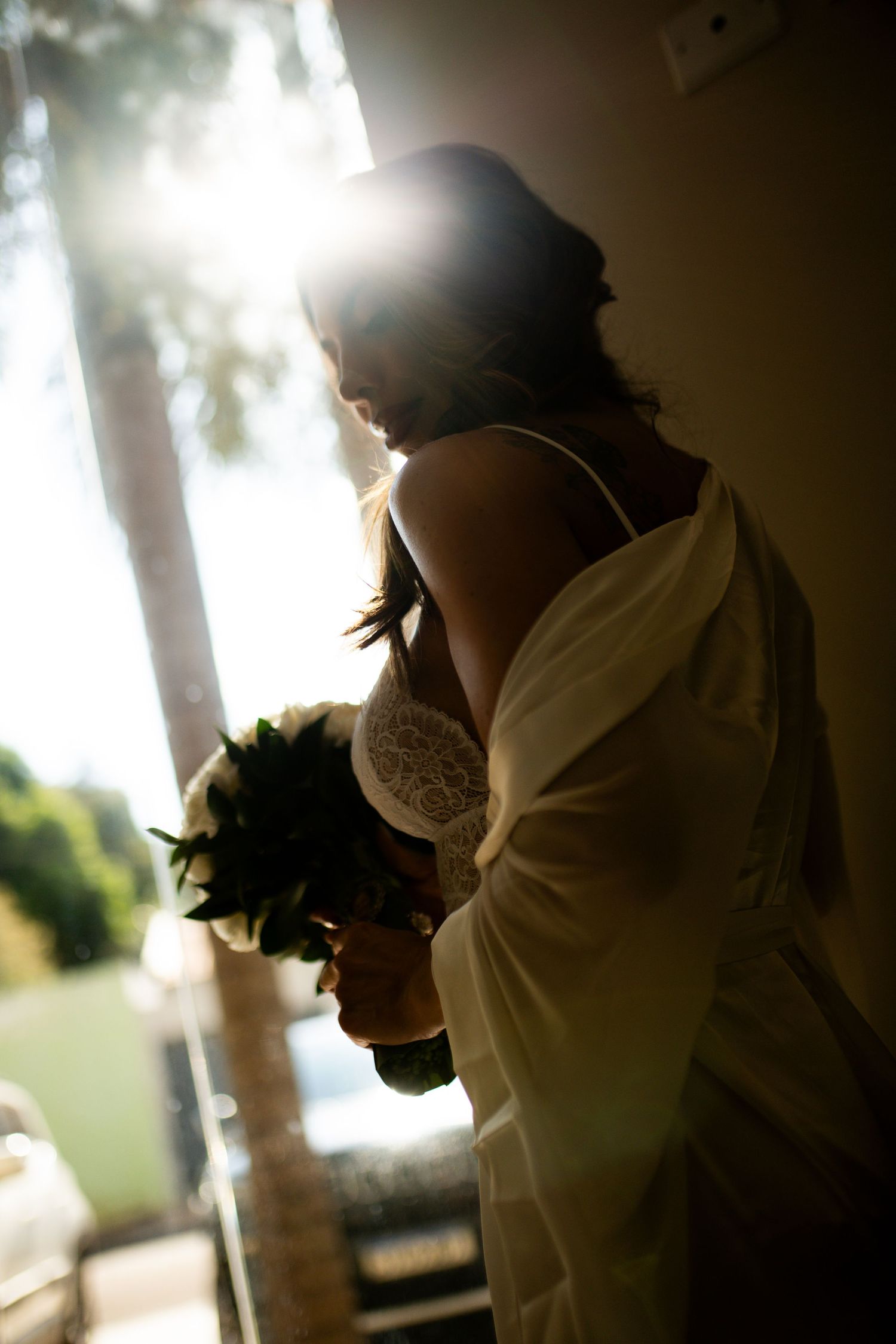 Silhouetted bride in white wedding dress holding bouquet standing by sunlit window.
