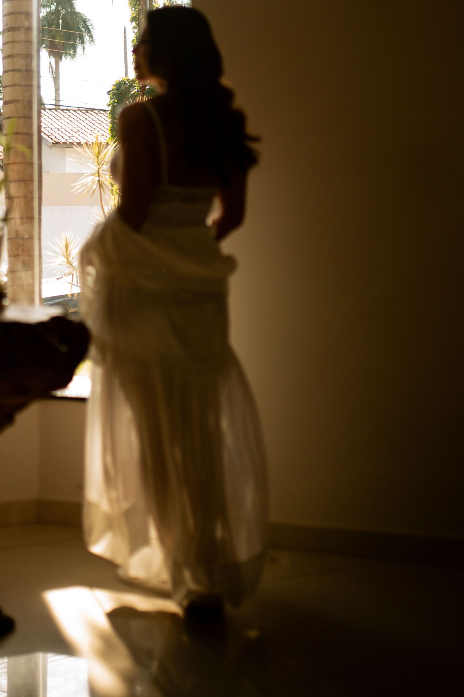 Silhouette of a woman in a flowing white dress standing by a sunlit window with palm trees visible outside.