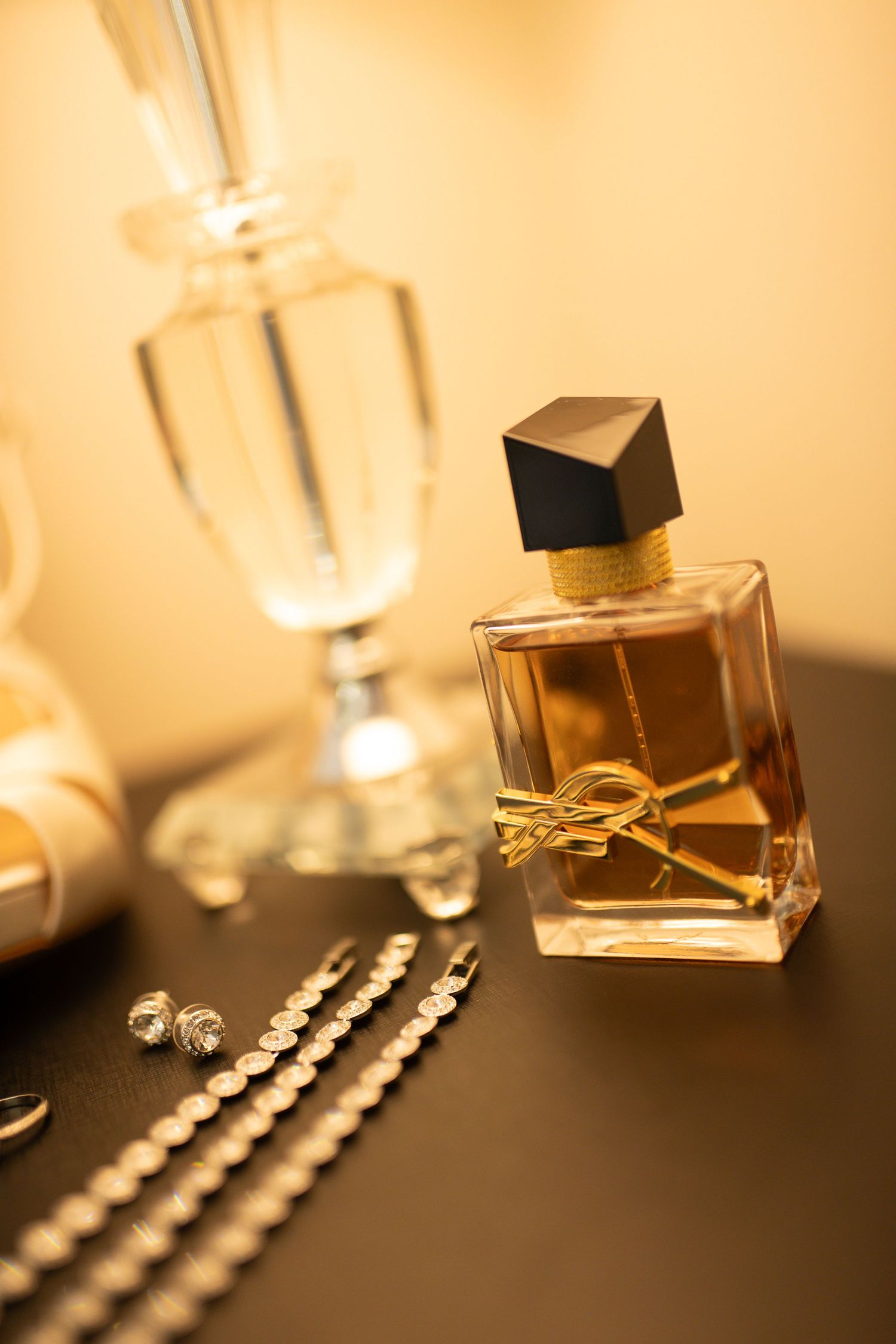 Elegant perfume bottle with gold bow and black cap beside crystal glassware and silver jewelry on dark surface.