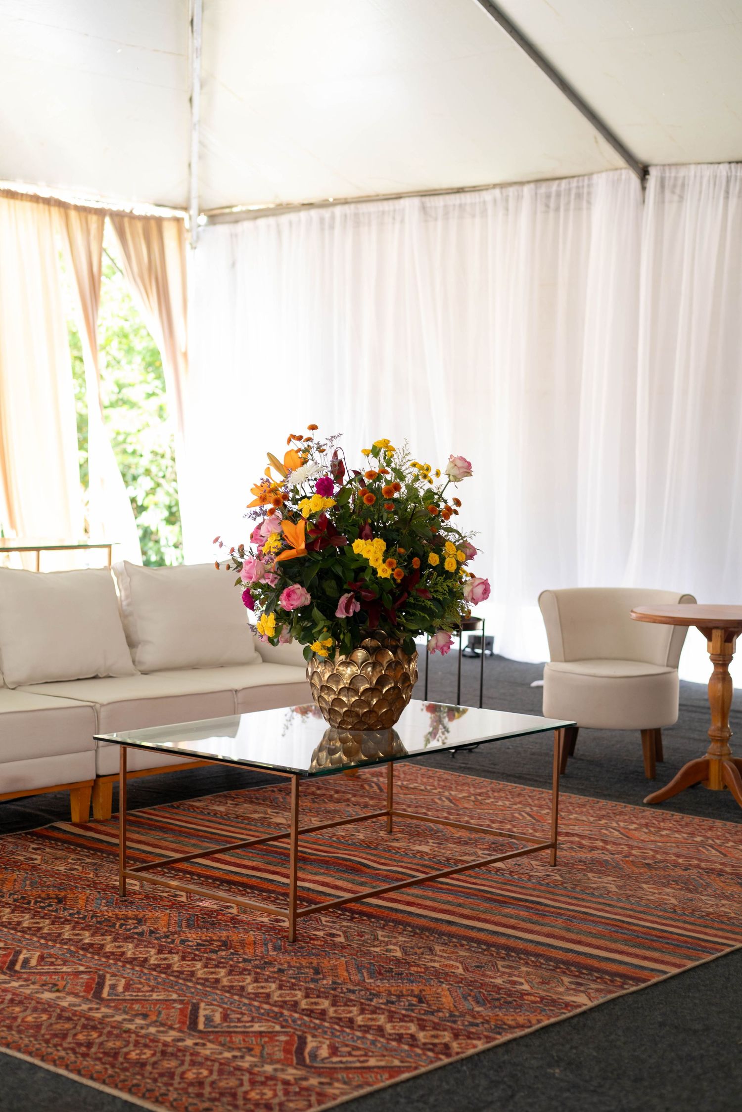 Elegant tent lounge with white sofa, gold coffee table, colorful floral arrangement, and patterned rug.
