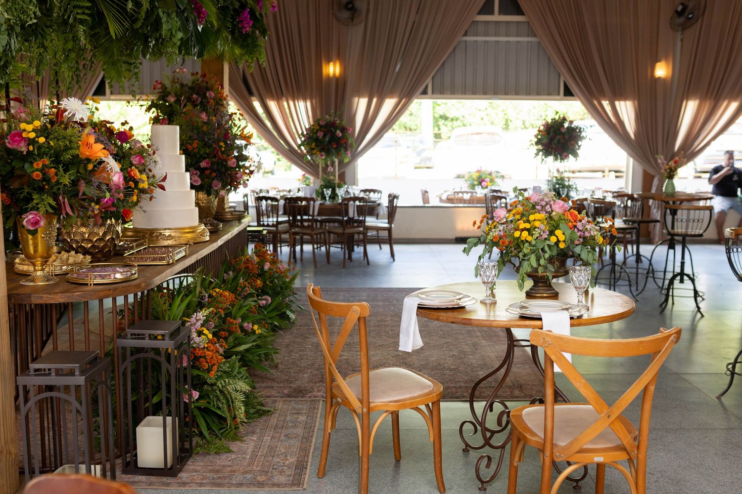 Elegant wedding reception venue with floral arrangements, wooden chairs, round tables, draped curtains, and a decorated cake table.