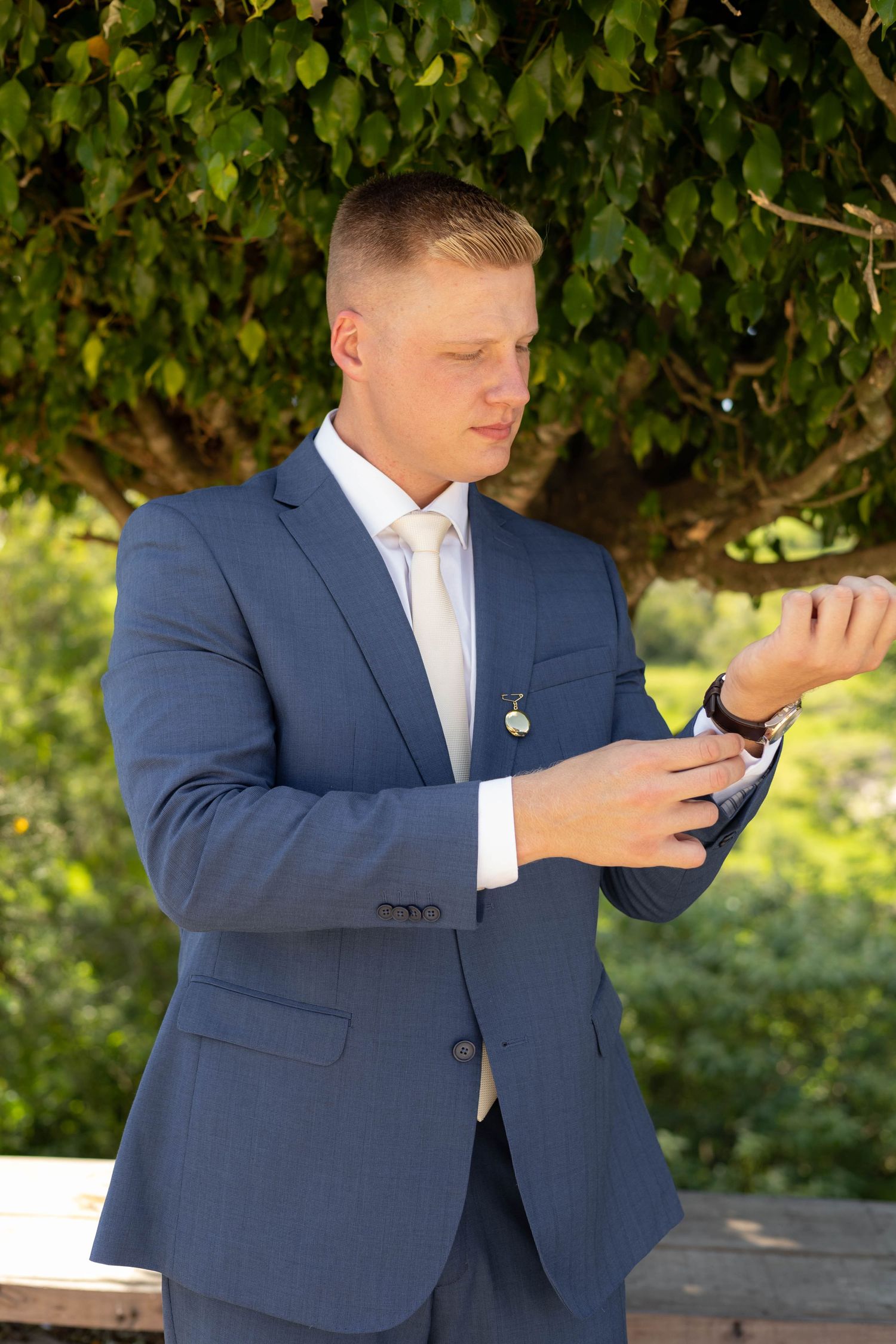 Young groom in blue suit adjusting cufflinks outdoors with lush green trees in background on wedding day.