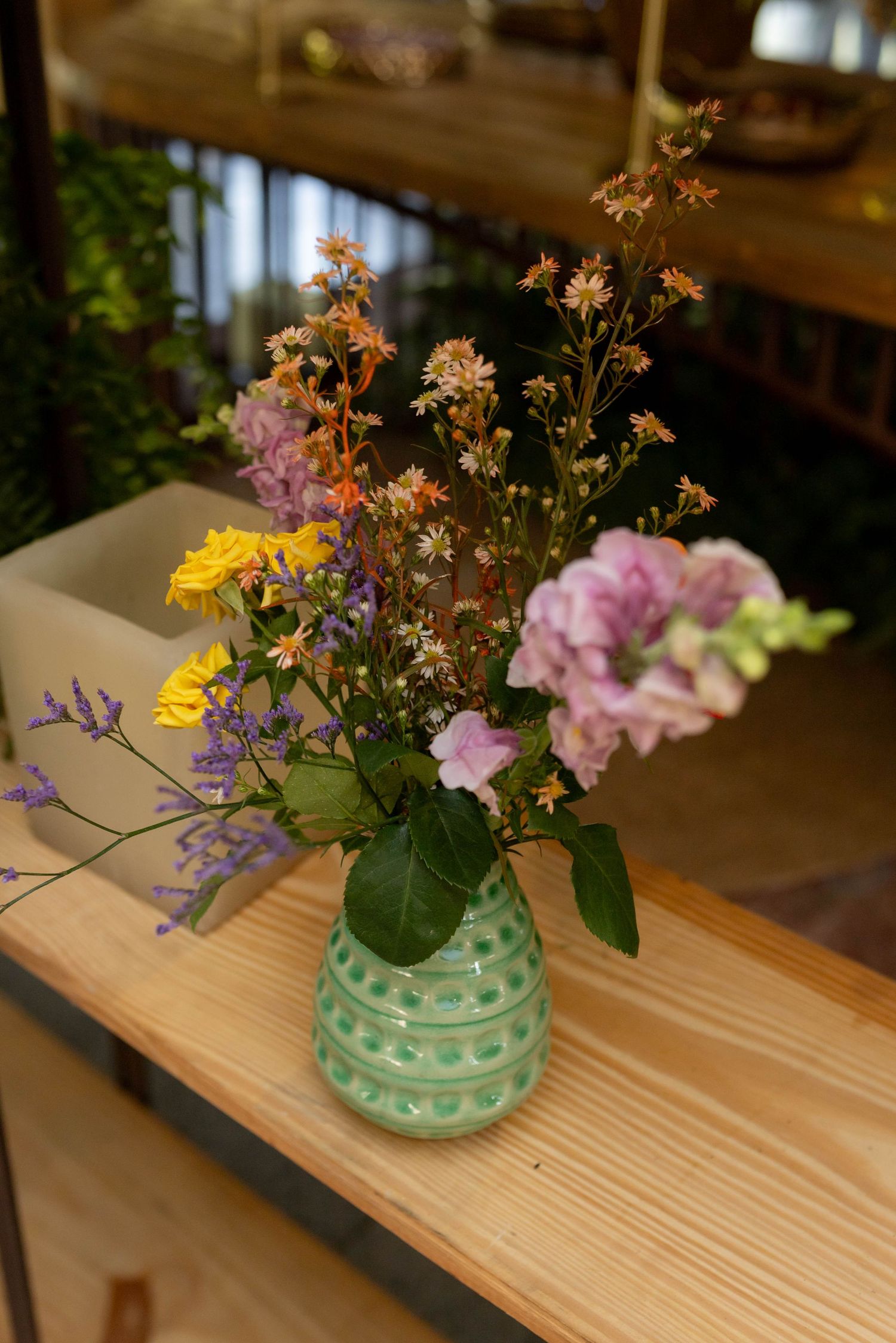 A green polka dot ceramic vase with colorful wildflowers including yellow, purple, and pink blooms on a wooden shelf.