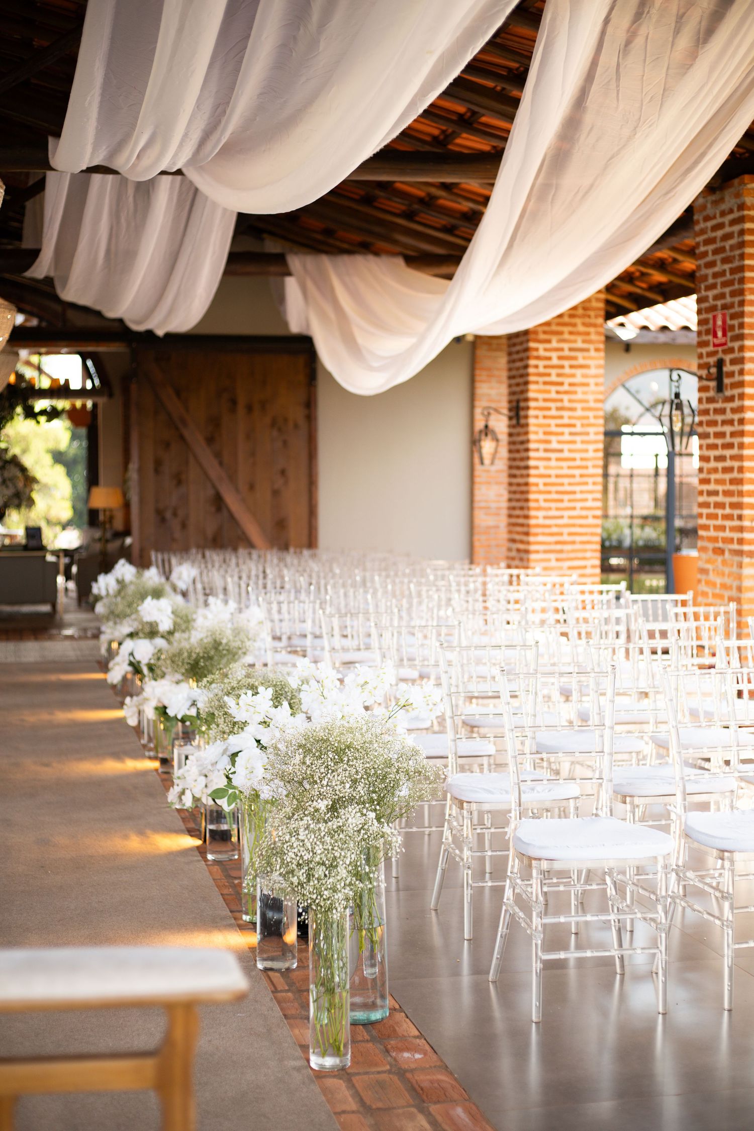 Elegant wedding ceremony aisle with white chiavari chairs, floral arrangements, and flowing white draping in a rustic brick venue.