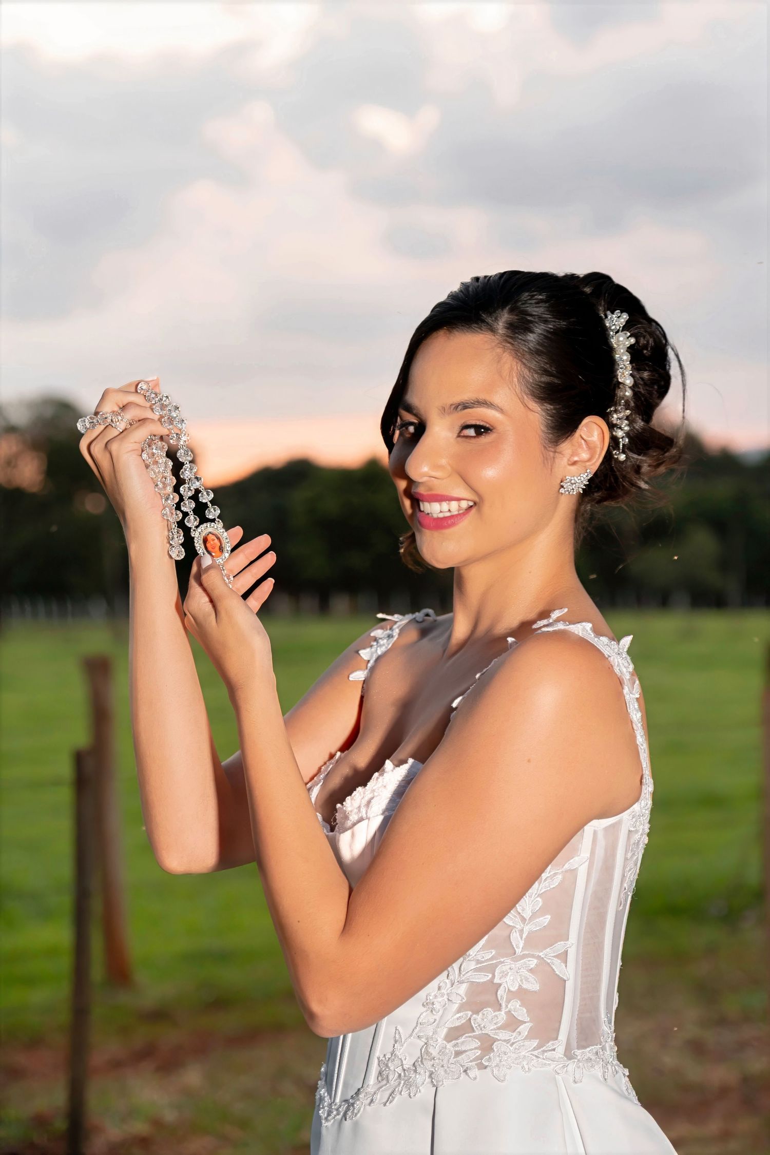 Bride in white lace gown holding crystal hair accessories, smiling outdoors at sunset with green fields behind her.