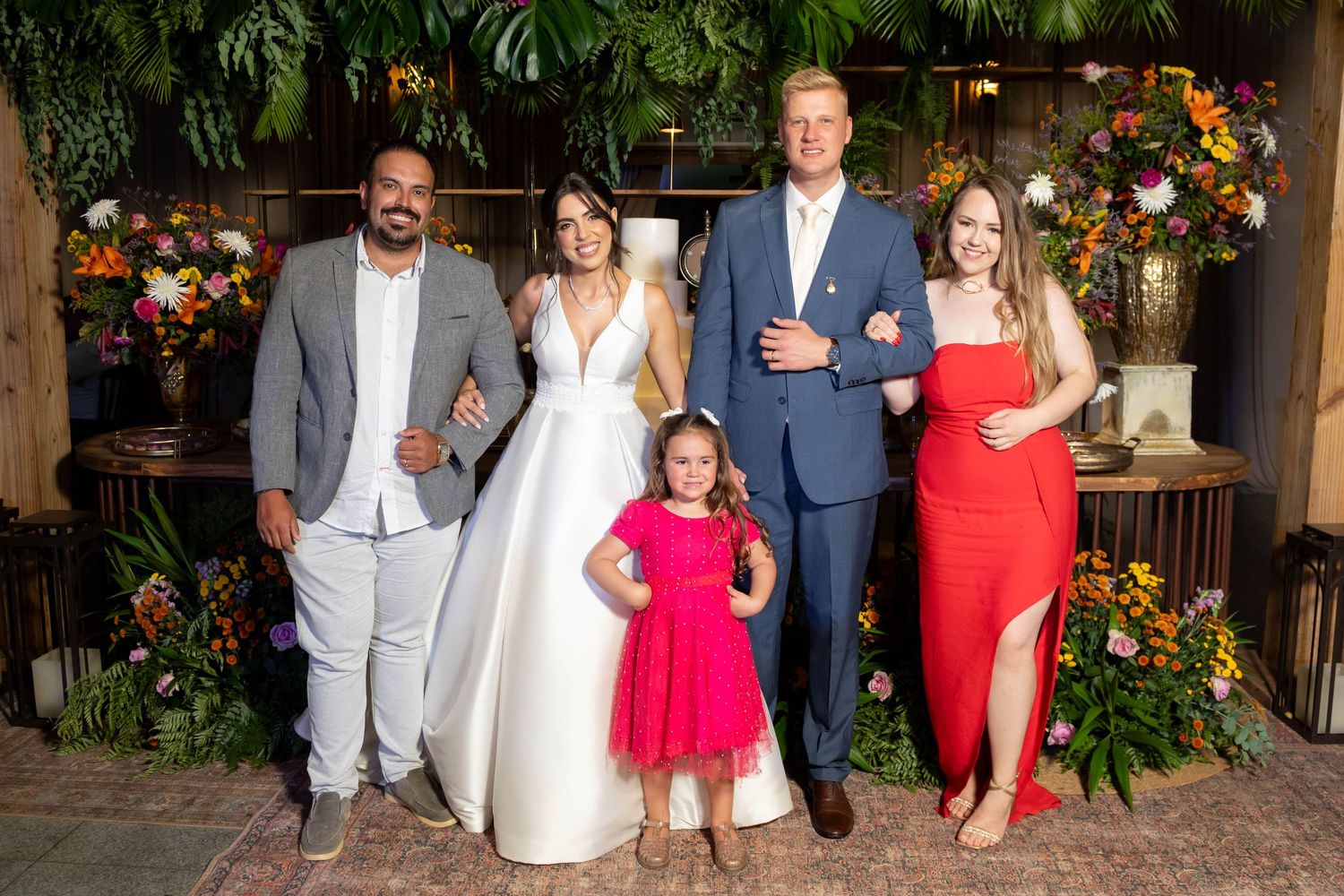 A joyful wedding group photo featuring a bride in white, groom in blue suit, and three guests amid lush floral decor.