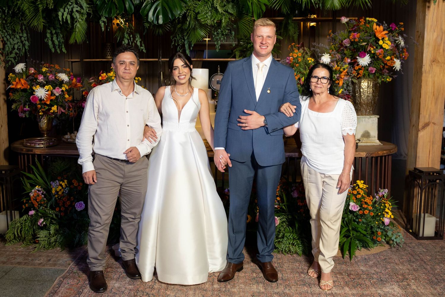 Bride and groom pose with two family members in front of lush floral backdrop at elegant wedding reception.