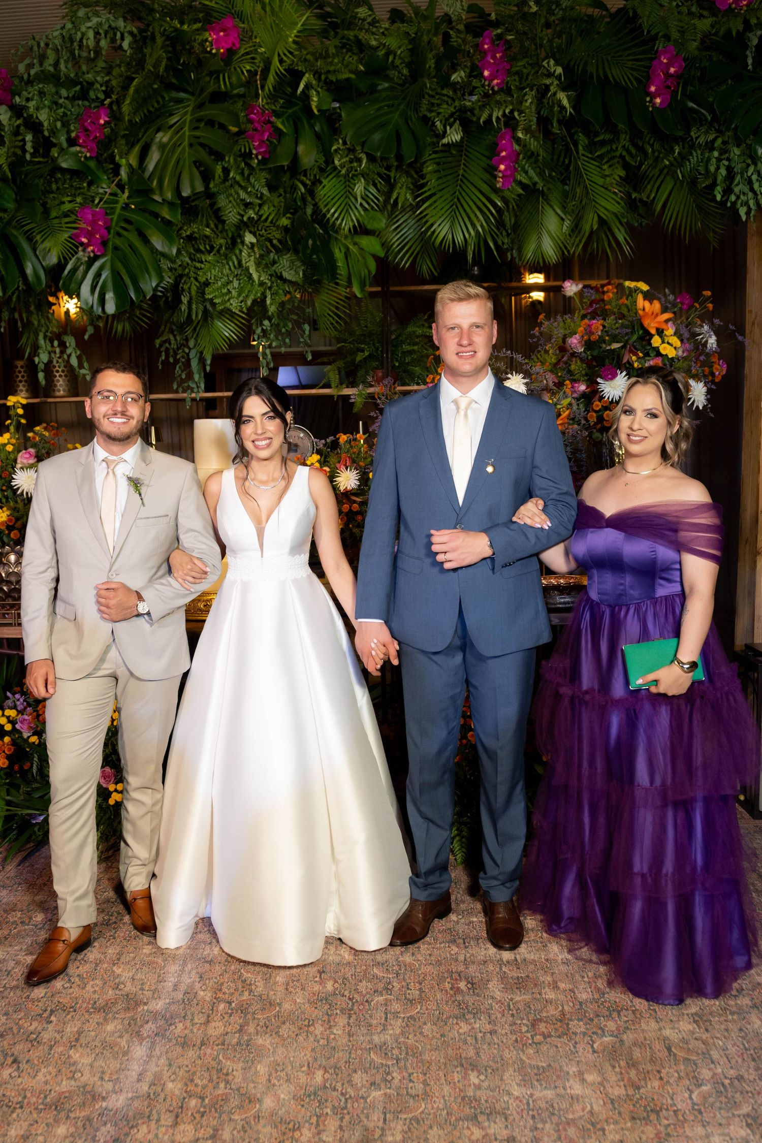 Wedding party of four posing indoors, bride in white gown, groom in blue suit, guests in purple dress and tan suit.