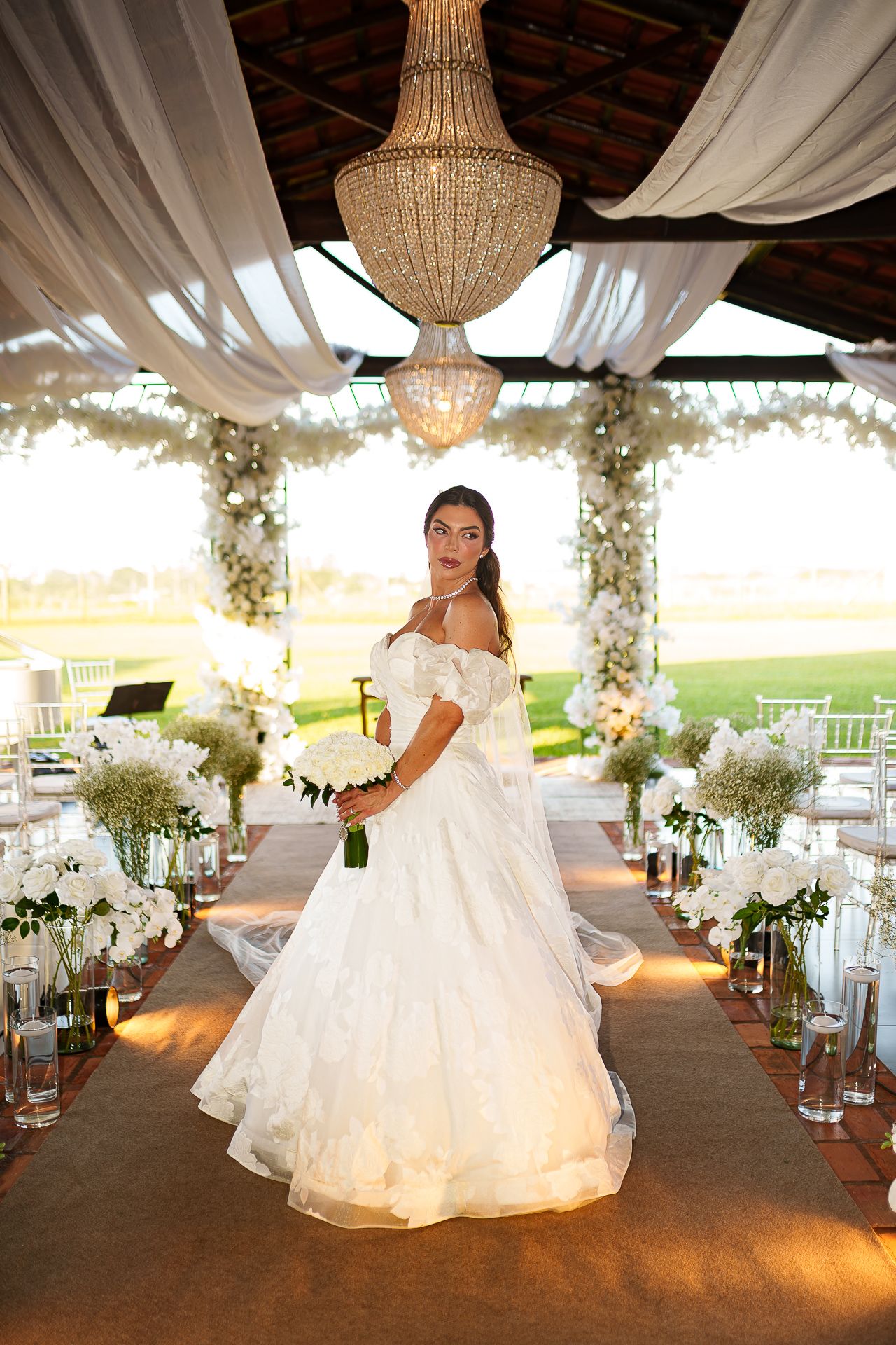 Bride in white off-shoulder ball gown holds bouquet at elegant floral wedding aisle with chandelier overhead.