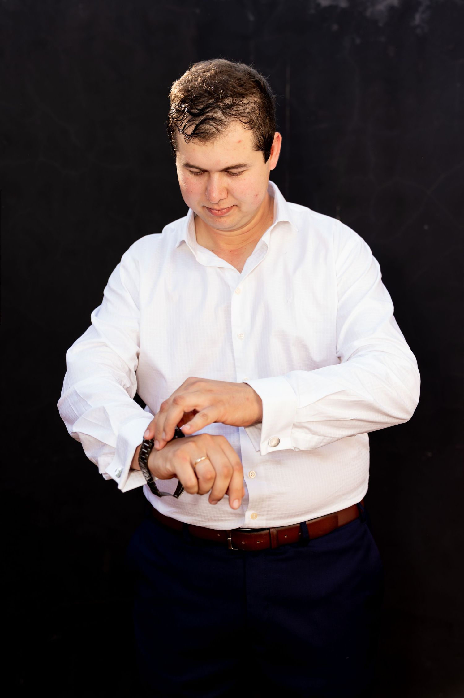 Man in white dress shirt adjusting his watch cufflinks against a dark background.