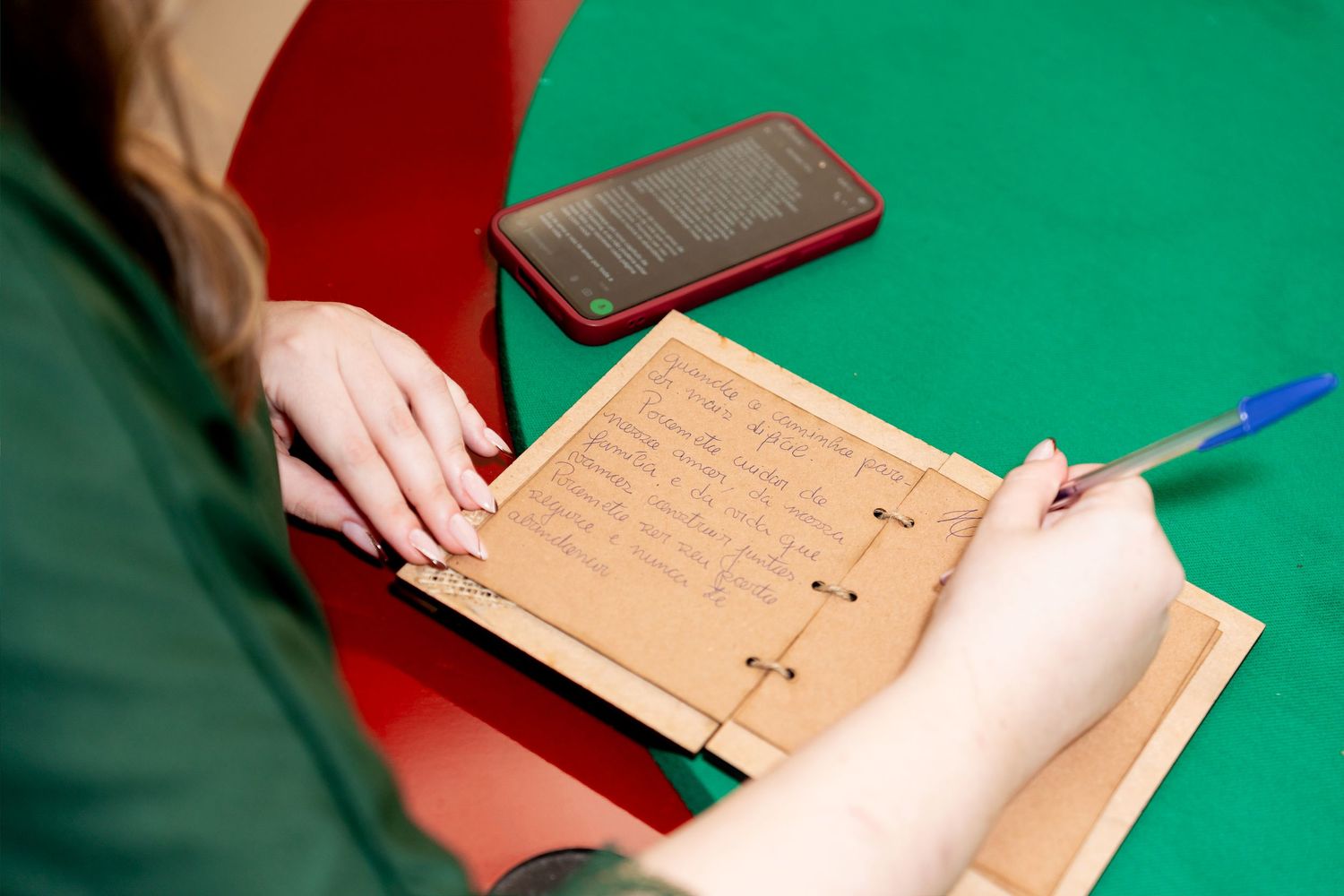 Person writing notes on a cardboard notebook with a blue pen on a green surface, with a smartphone nearby.