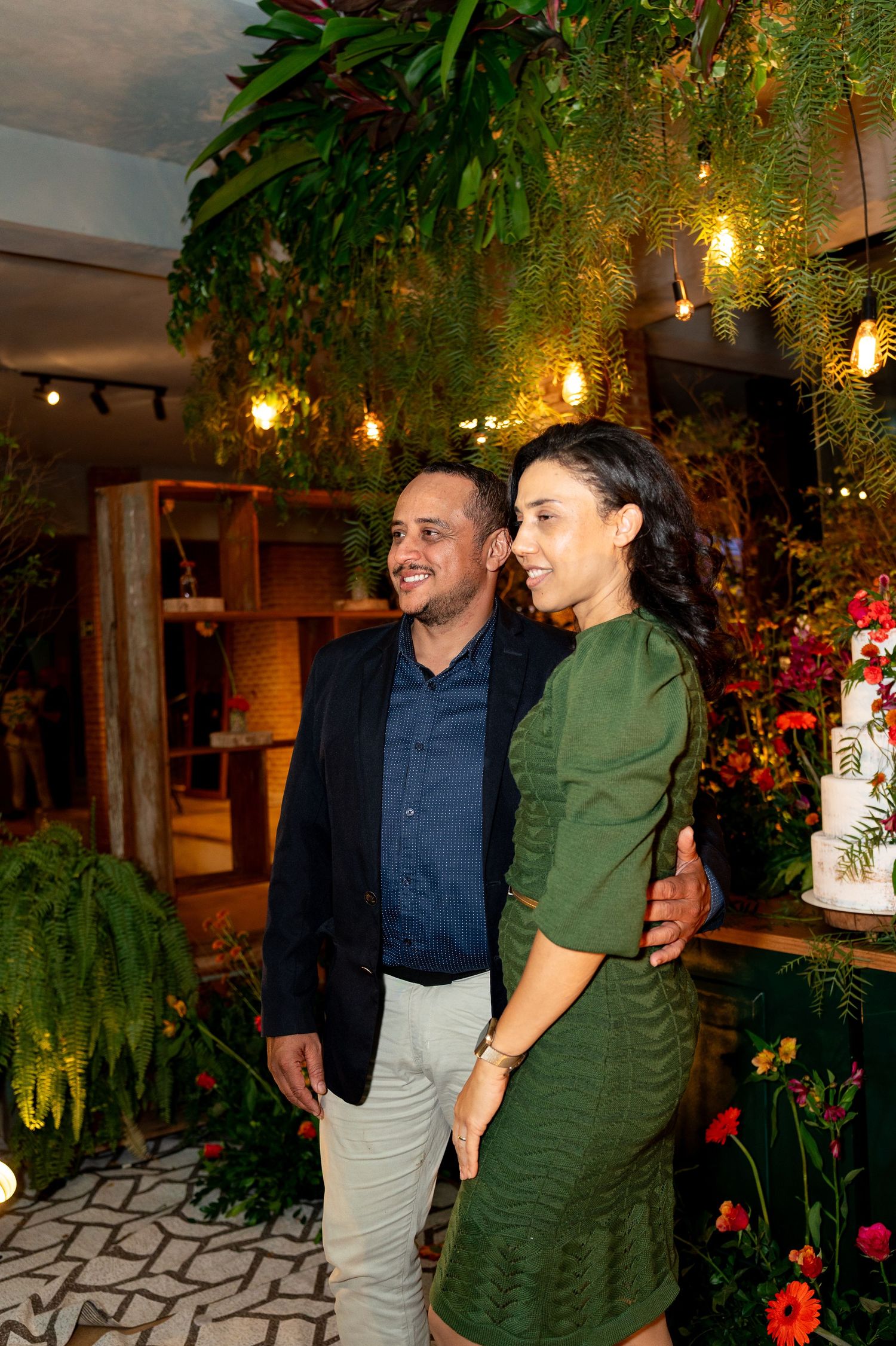 Couple smiling and embracing at an elegant event with lush greenery, warm lights, and a white cake in the background.