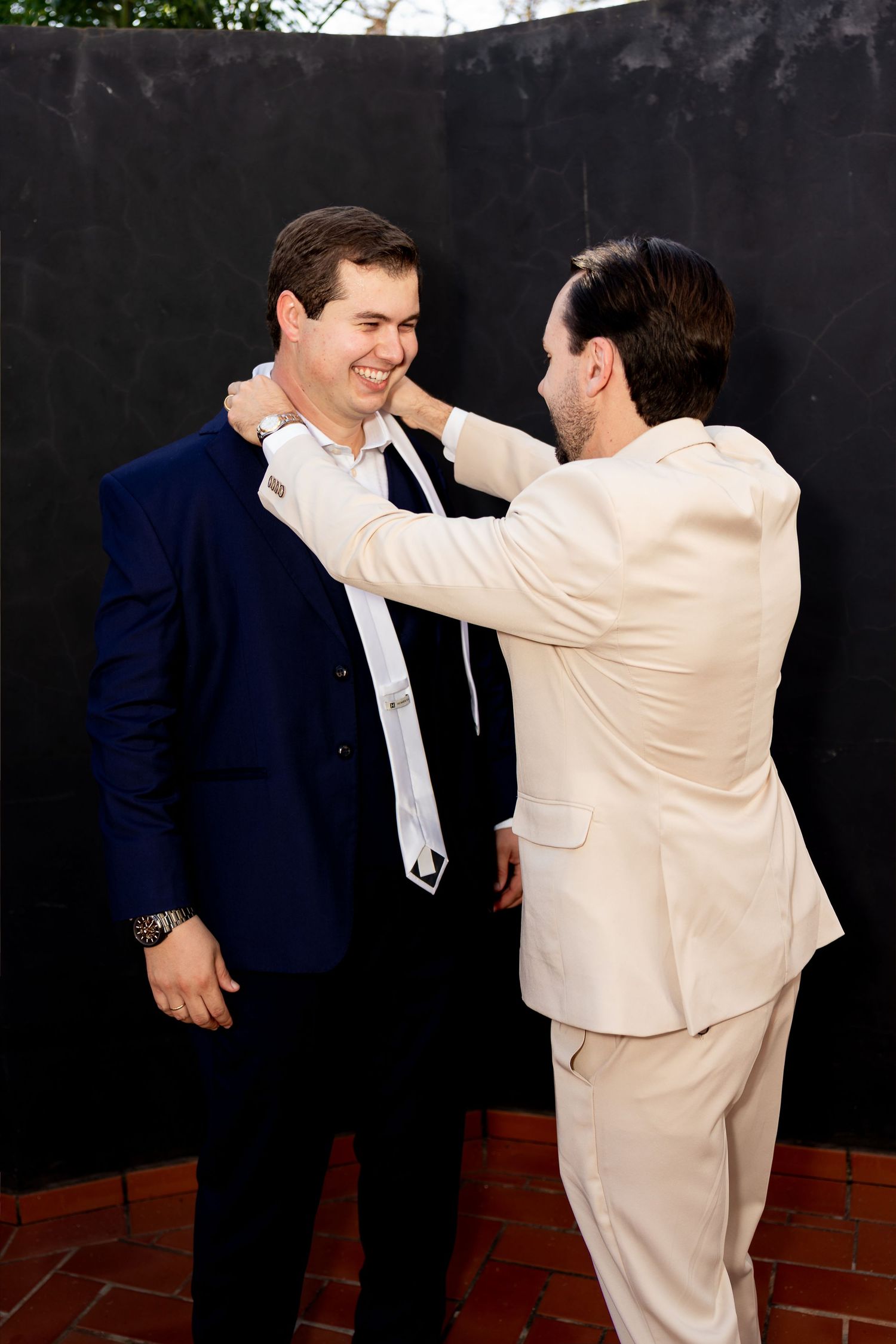 Two men in suits, one in navy and one in cream, adjusting a white tie before a formal event.