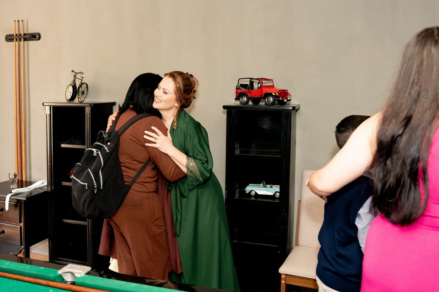 Two women hugging warmly in a room with a pool table, while others watch nearby.