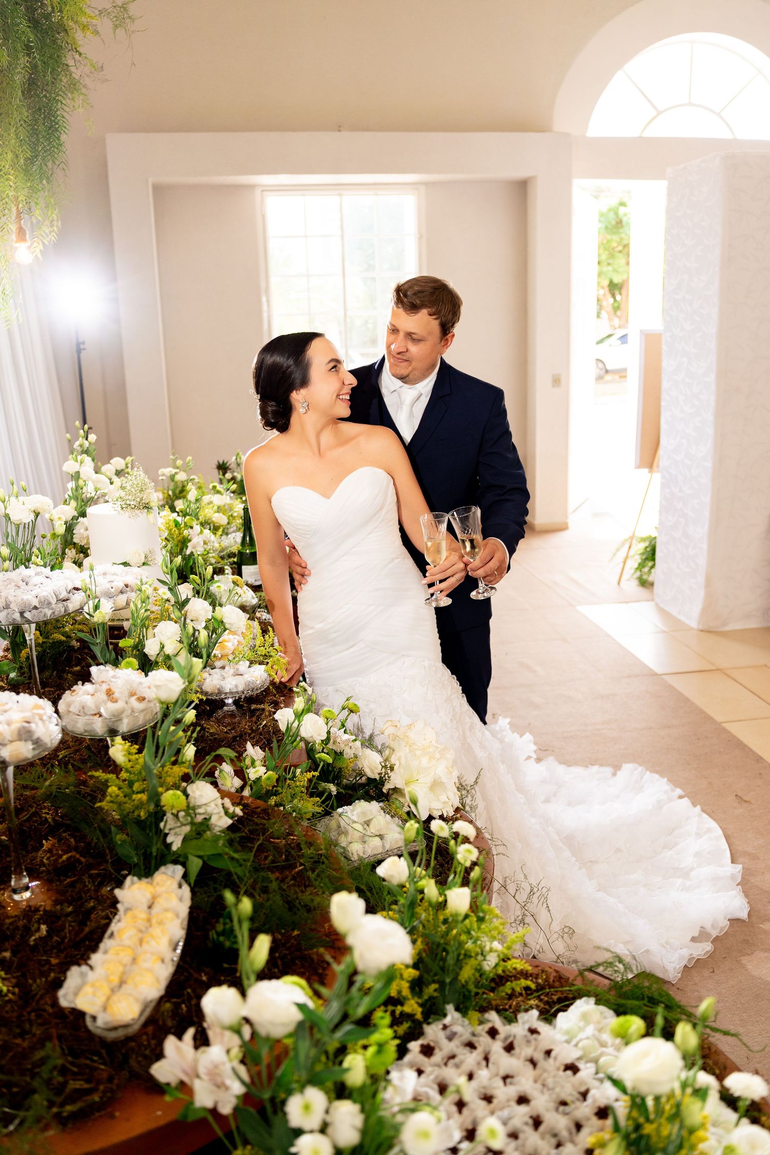 Bride and groom toasting with champagne glasses beside white floral arrangements at elegant wedding reception.