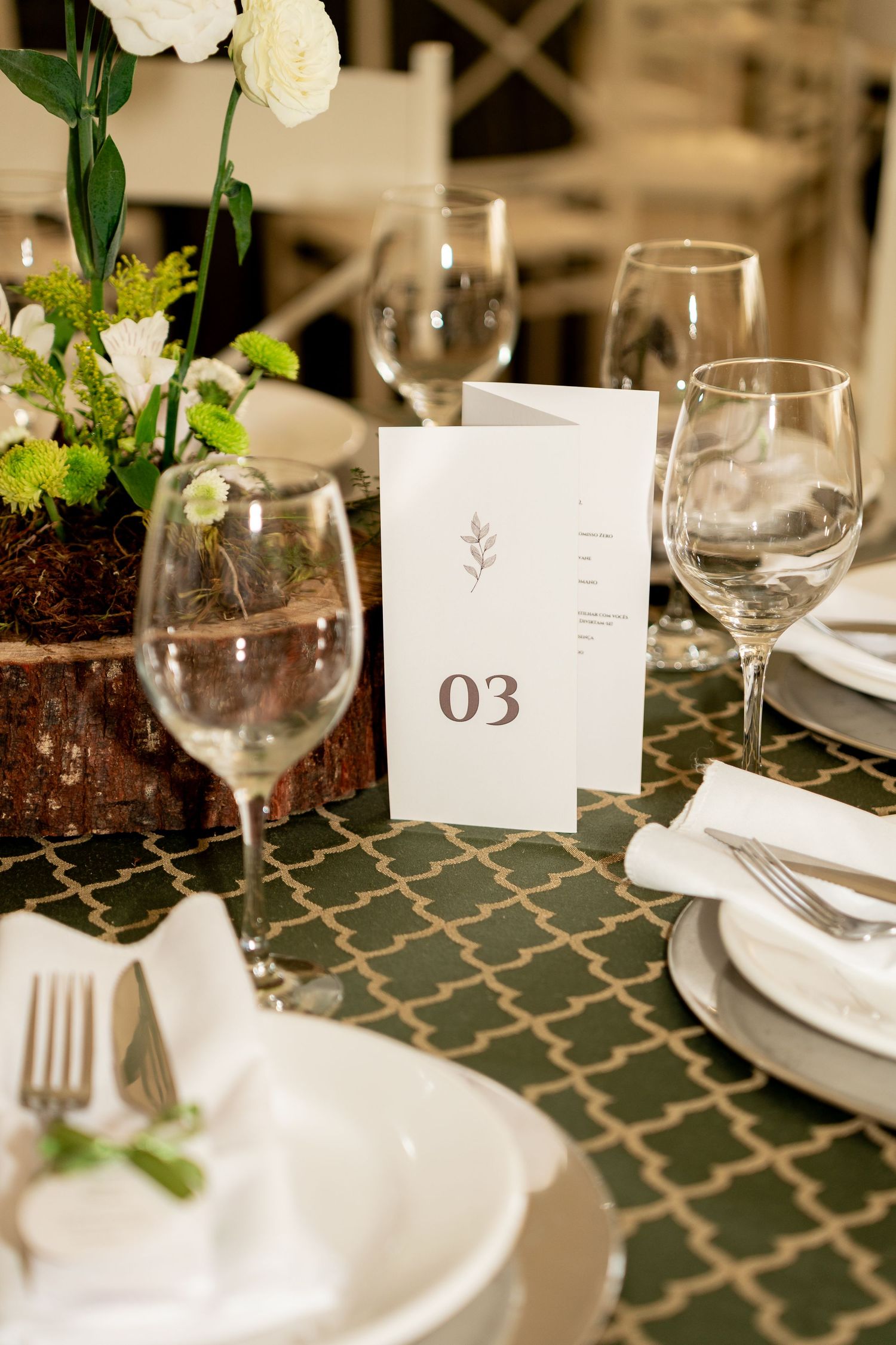 Elegant wedding table setting with wine glasses, white plates, floral centerpiece, and table number 03 card on green tablecloth.