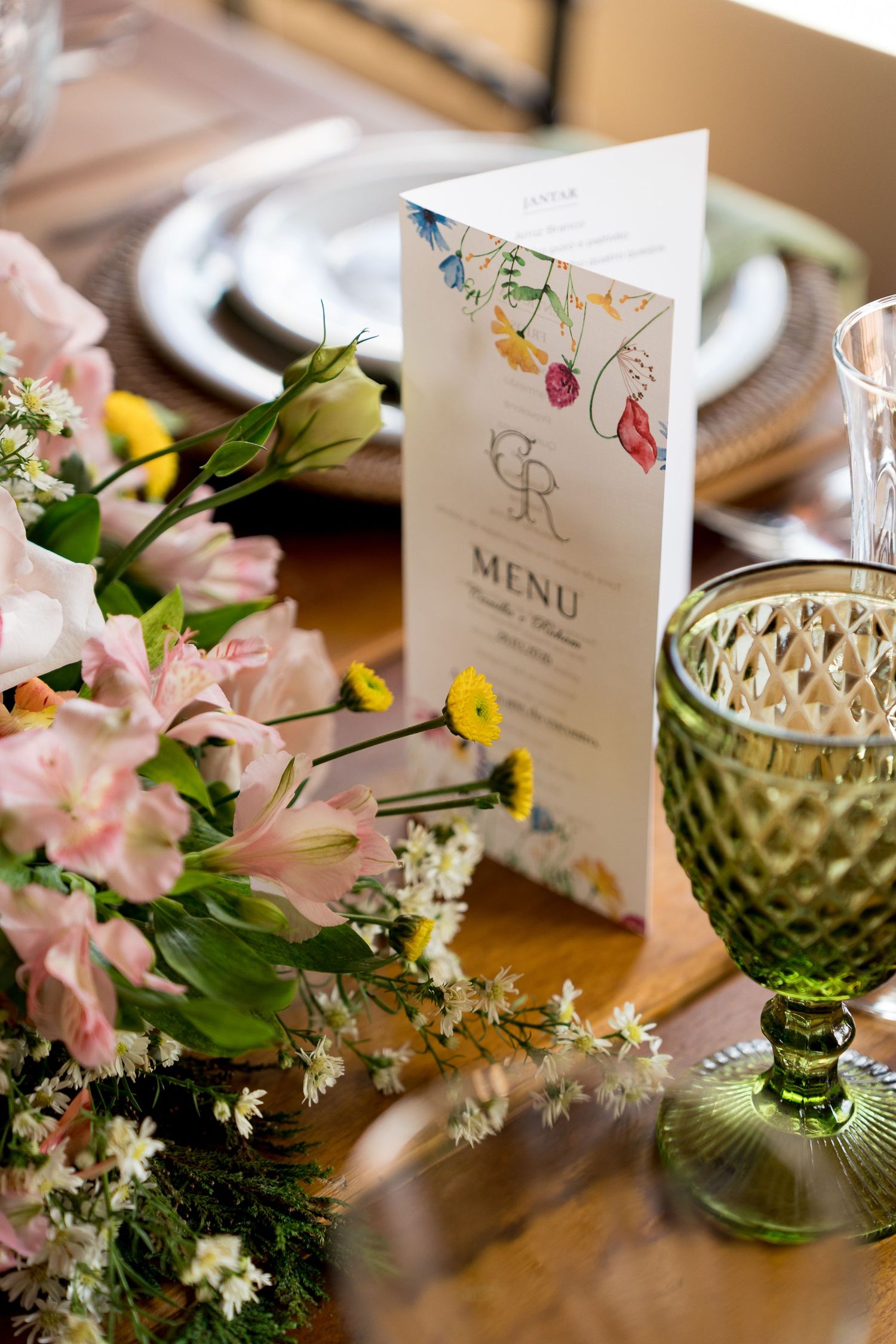 Elegant wedding table setting with pink flowers, green glassware, and a decorative menu card with floral accents on a wooden table.