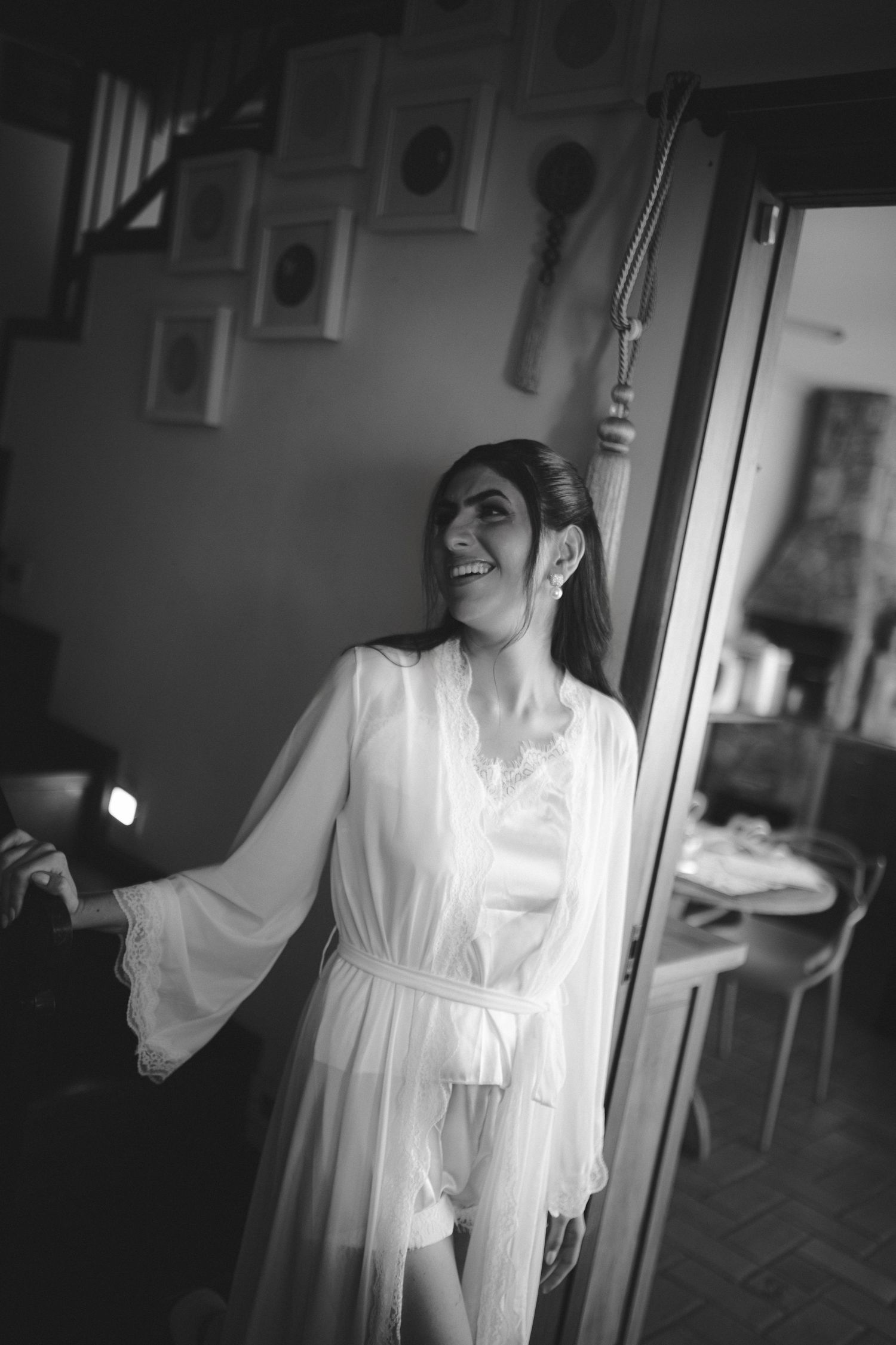Black and white photo of a smiling bride in a white lace robe standing in a doorway during wedding preparations.