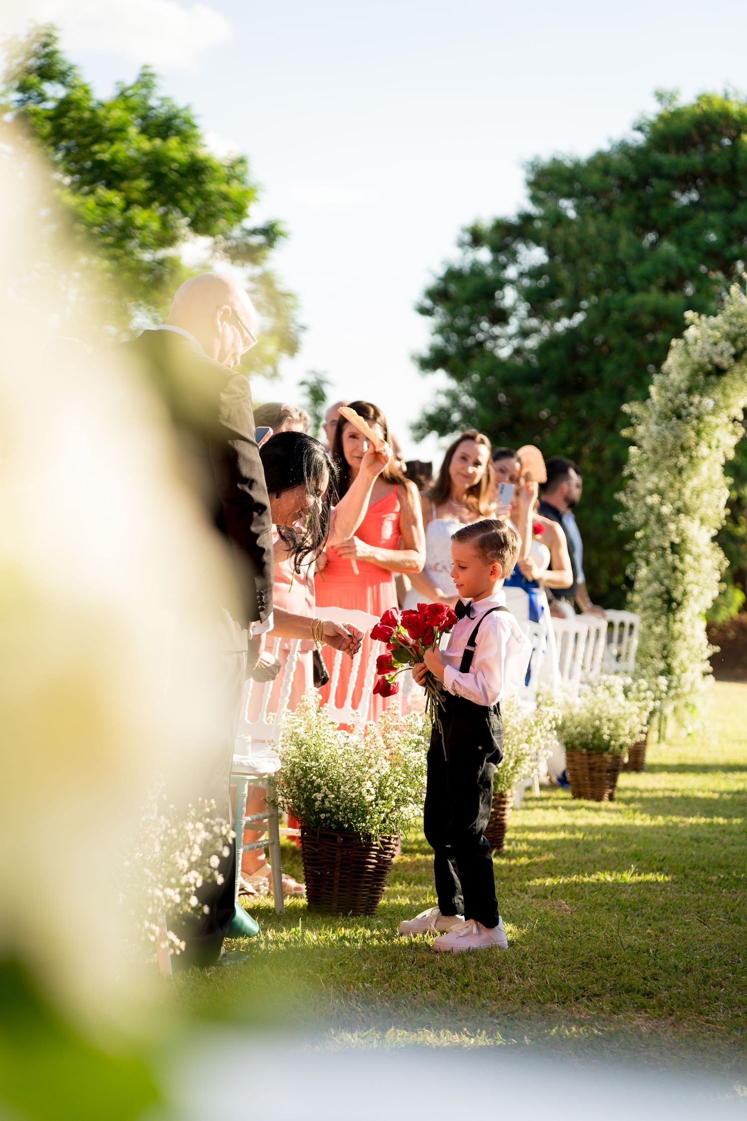Young ring bearer holding red flowers walks down outdoor wedding aisle while guests applaud in sunny garden ceremony.