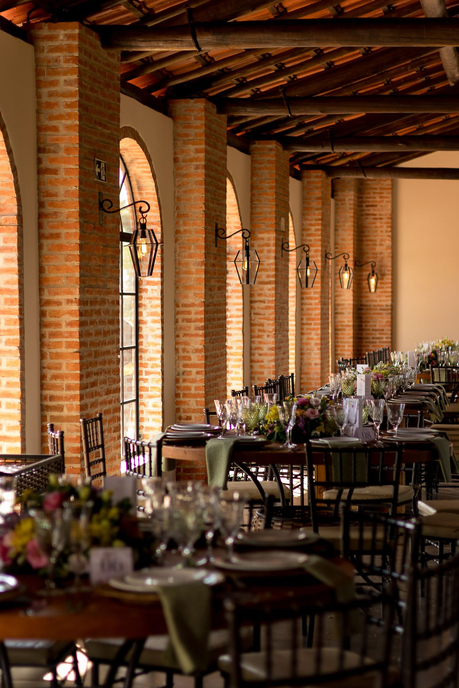 Elegant rustic wedding reception venue with brick columns, arched windows, hanging lanterns, and long tables set with floral centerpieces.