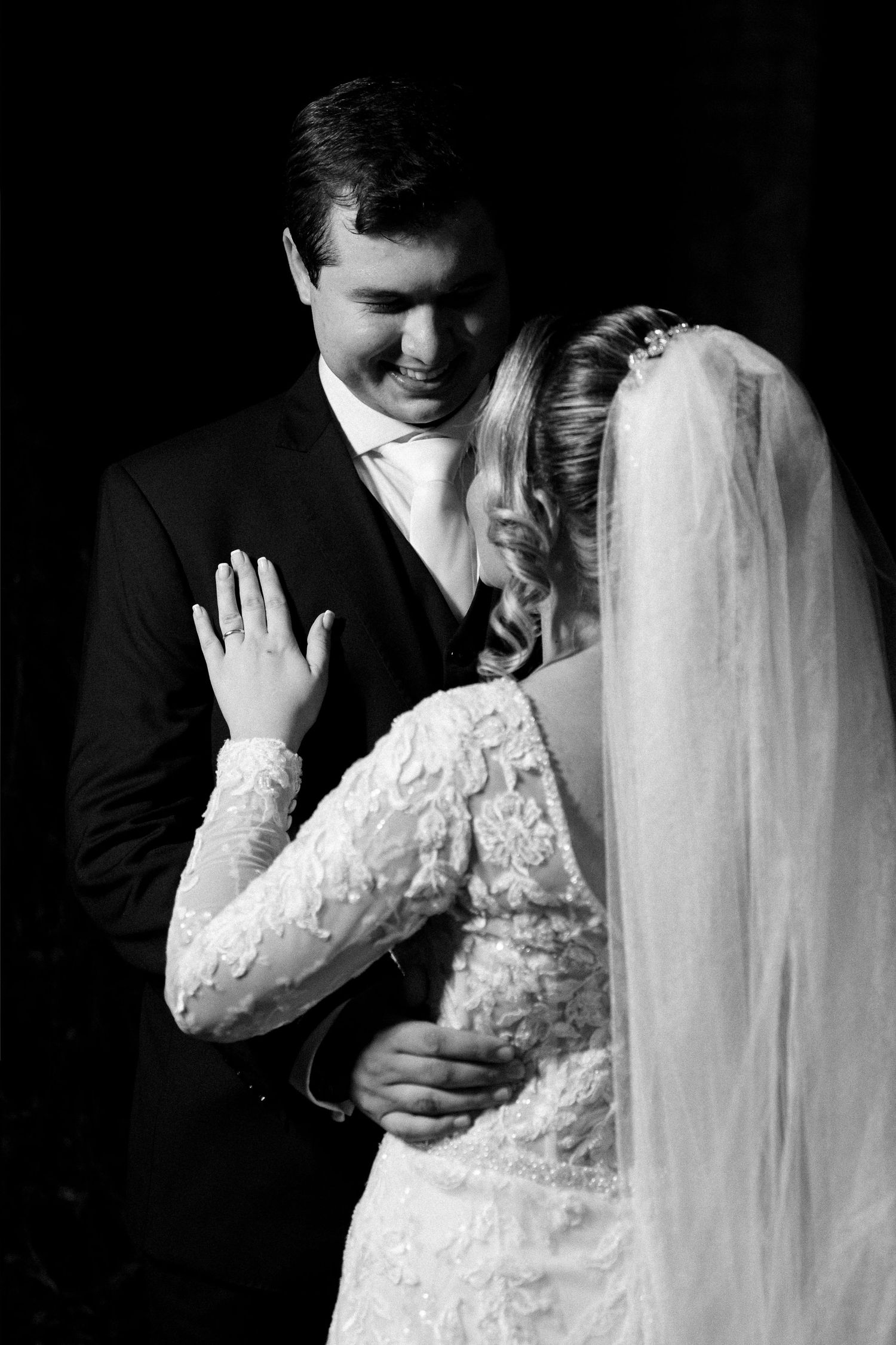 Black and white wedding photo of smiling groom in suit embracing bride in lace dress and veil.