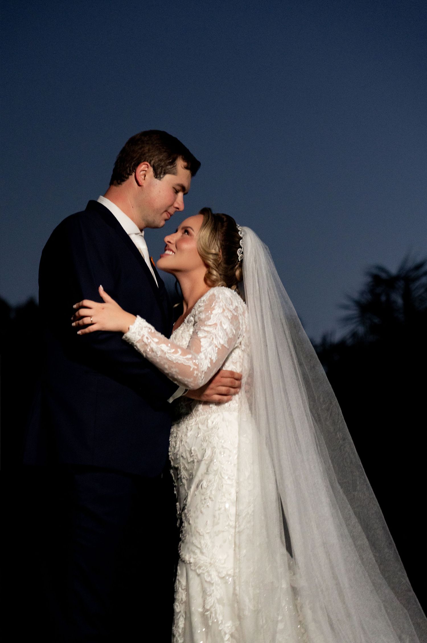 Bride and groom embrace romantically at dusk, her lace long-sleeve gown and veil glowing against the deep blue night sky.