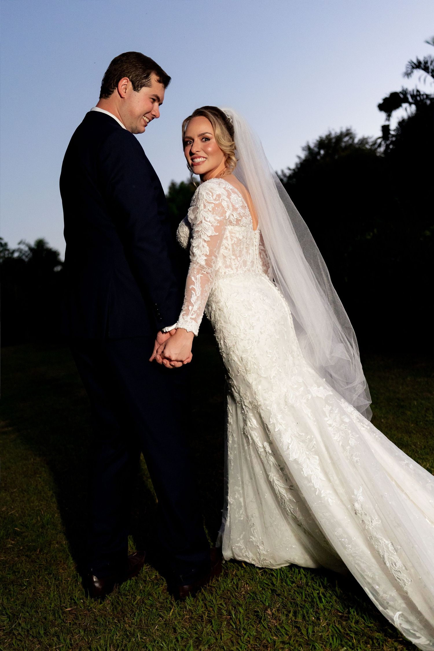Bride in lace mermaid gown with veil holds hands with groom in navy suit outdoors at golden hour.