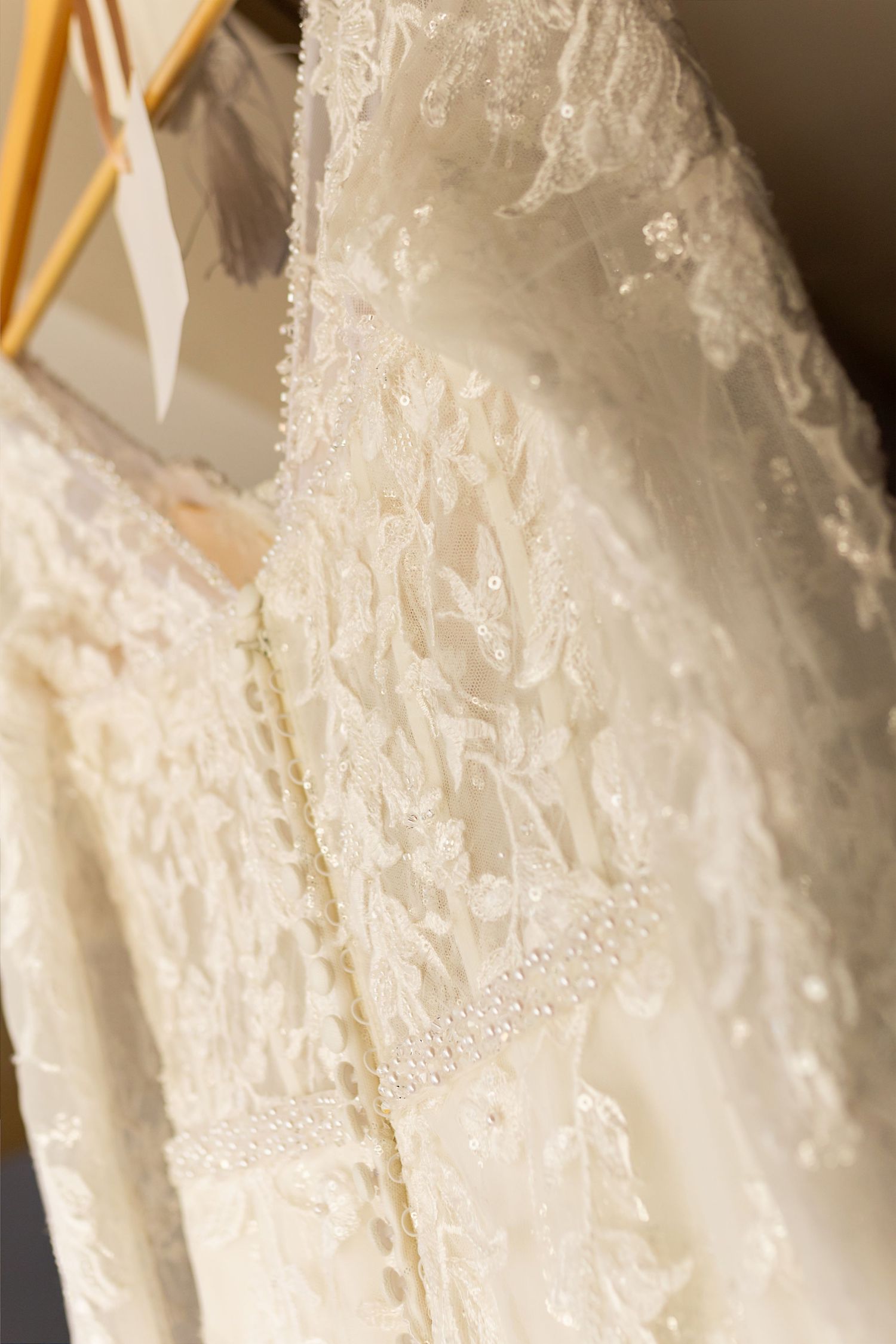 Close-up of an ivory lace wedding dress with pearl trim details hanging on gold hangers.
