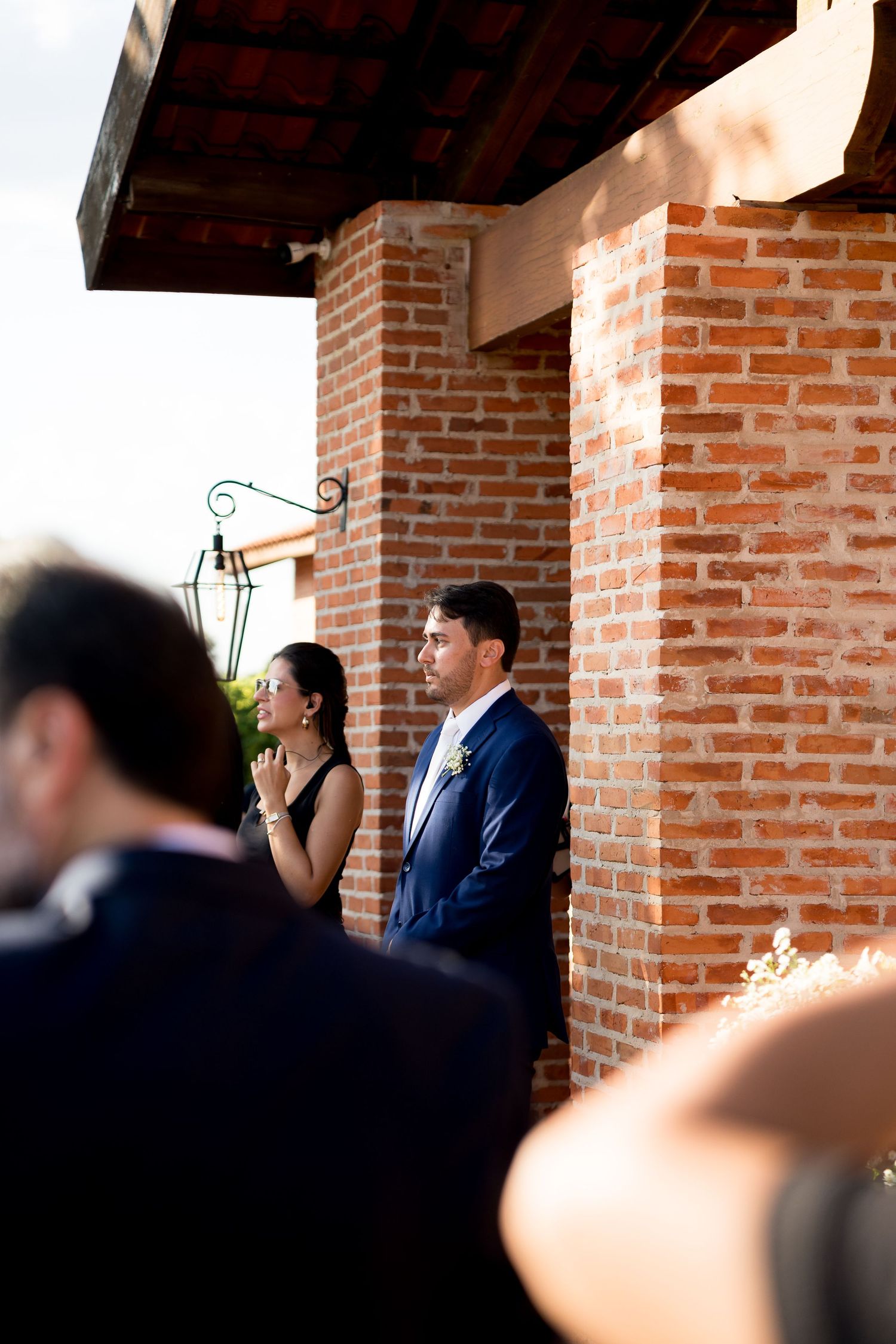 A groom in a blue suit stands with a woman in black dress against a brick wall at an outdoor wedding venue.