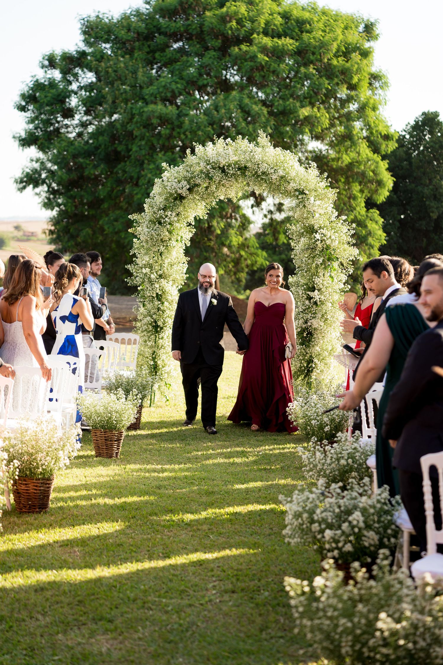 Couple walks down outdoor wedding aisle under white floral arch, bride in burgundy gown, guests watching on green lawn.