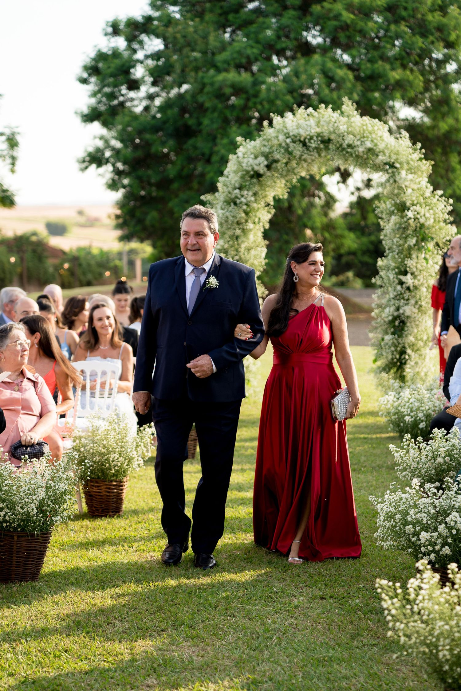 Man in navy suit escorts woman in red gown down floral arch wedding aisle outdoors.