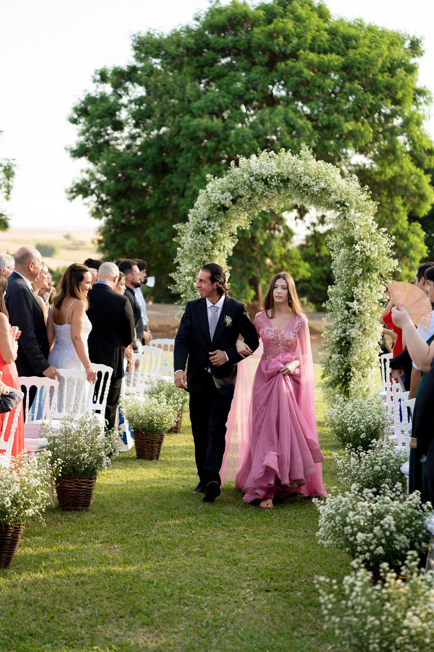 Couple walks down floral arch aisle at outdoor wedding ceremony surrounded by white flower arrangements.