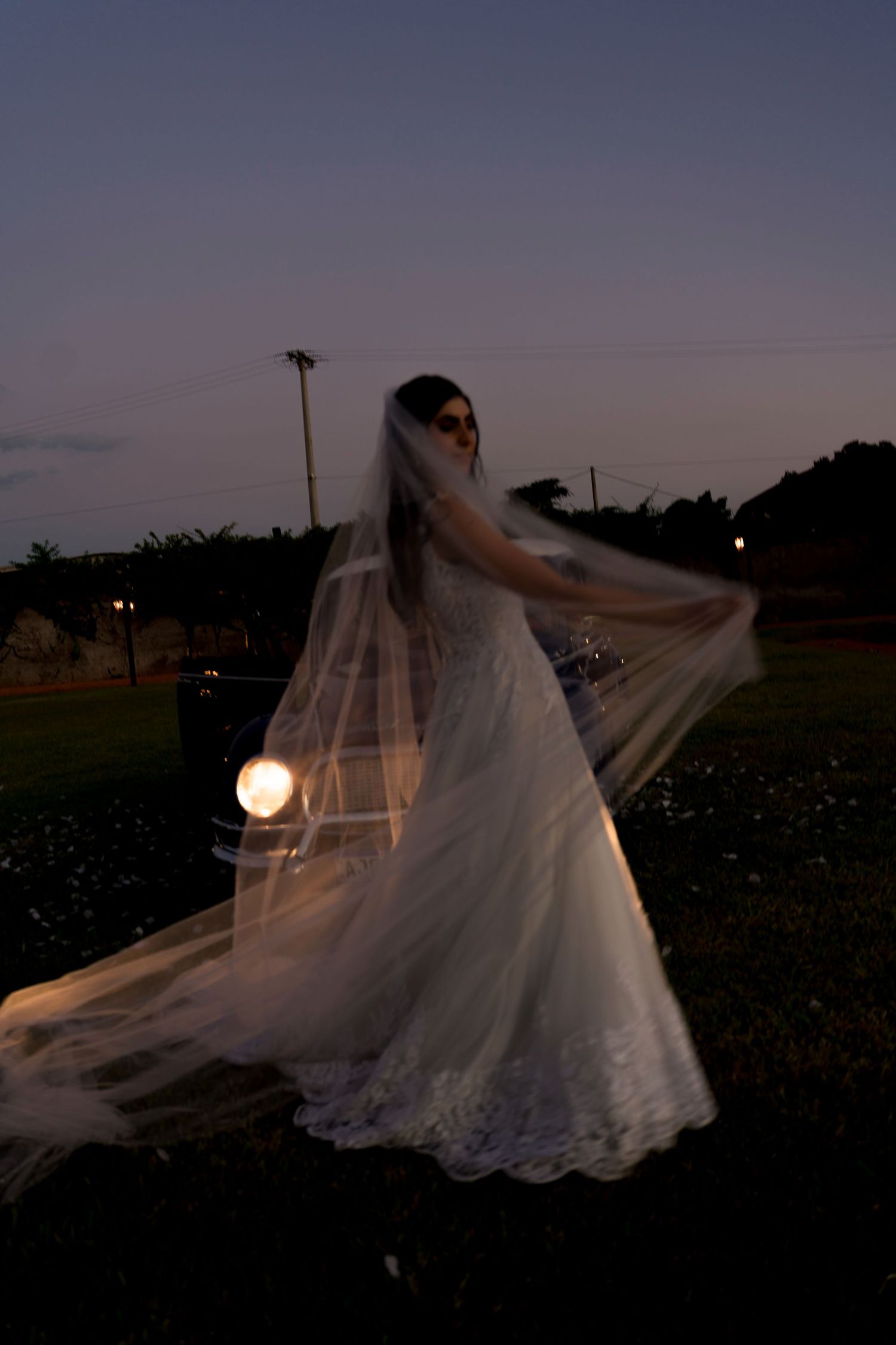 Bride in white wedding gown and veil posing outdoors at dusk with soft light glowing behind her.