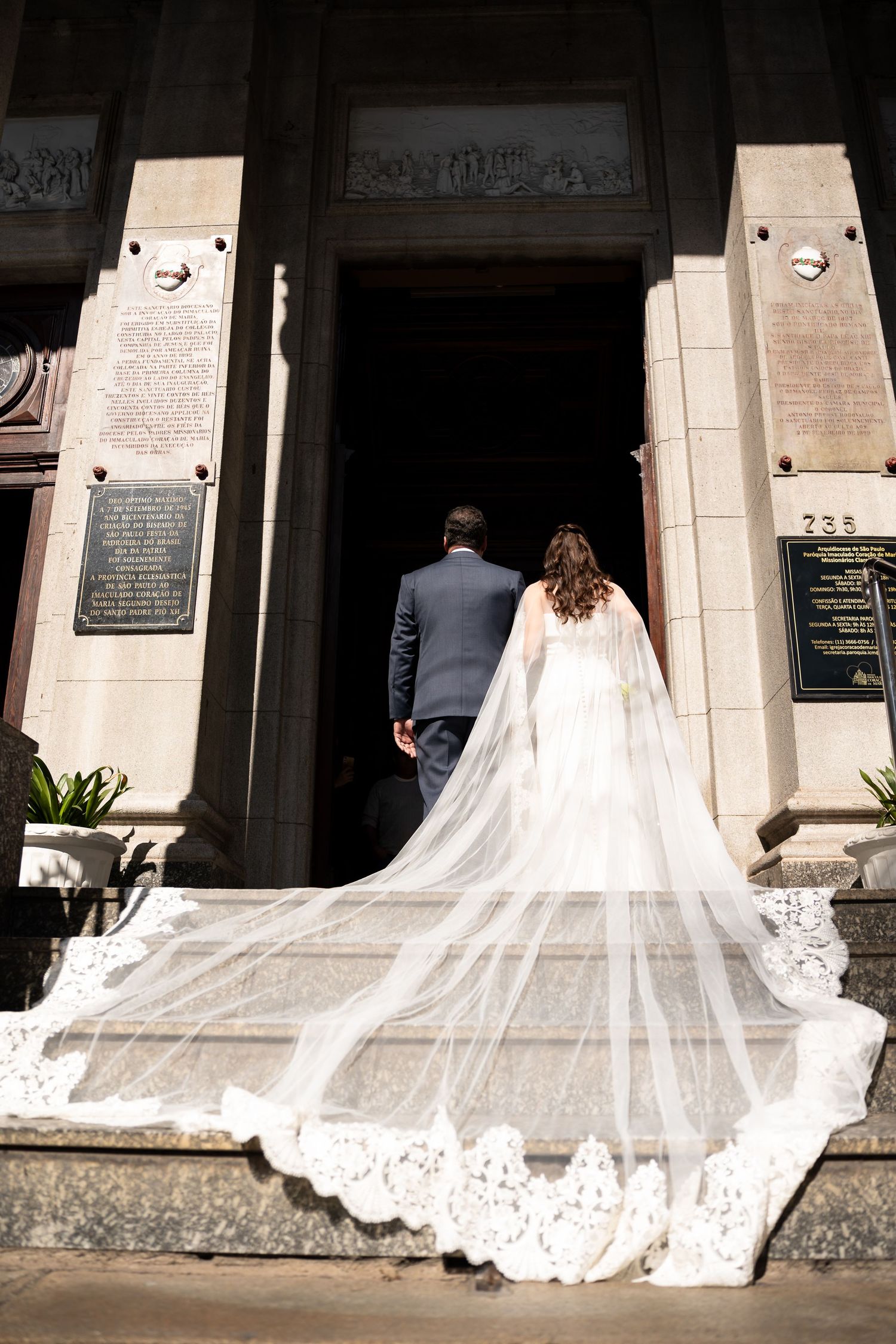 Bride and groom walking into church entrance, long lace cathedral veil trailing down stone steps on wedding day.