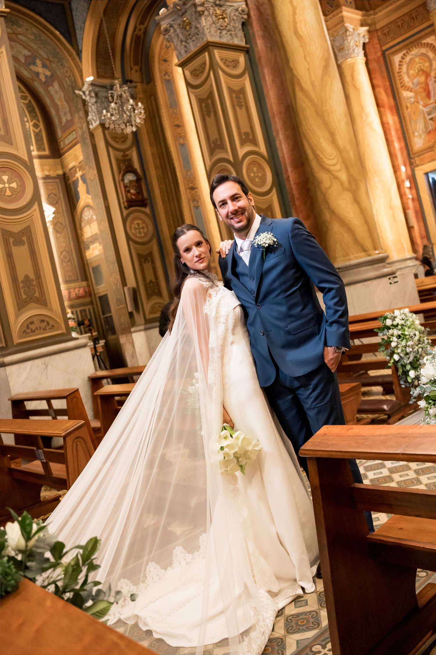 A smiling bride and groom pose together inside an ornate church, she in a white gown with cape, he in a navy suit.