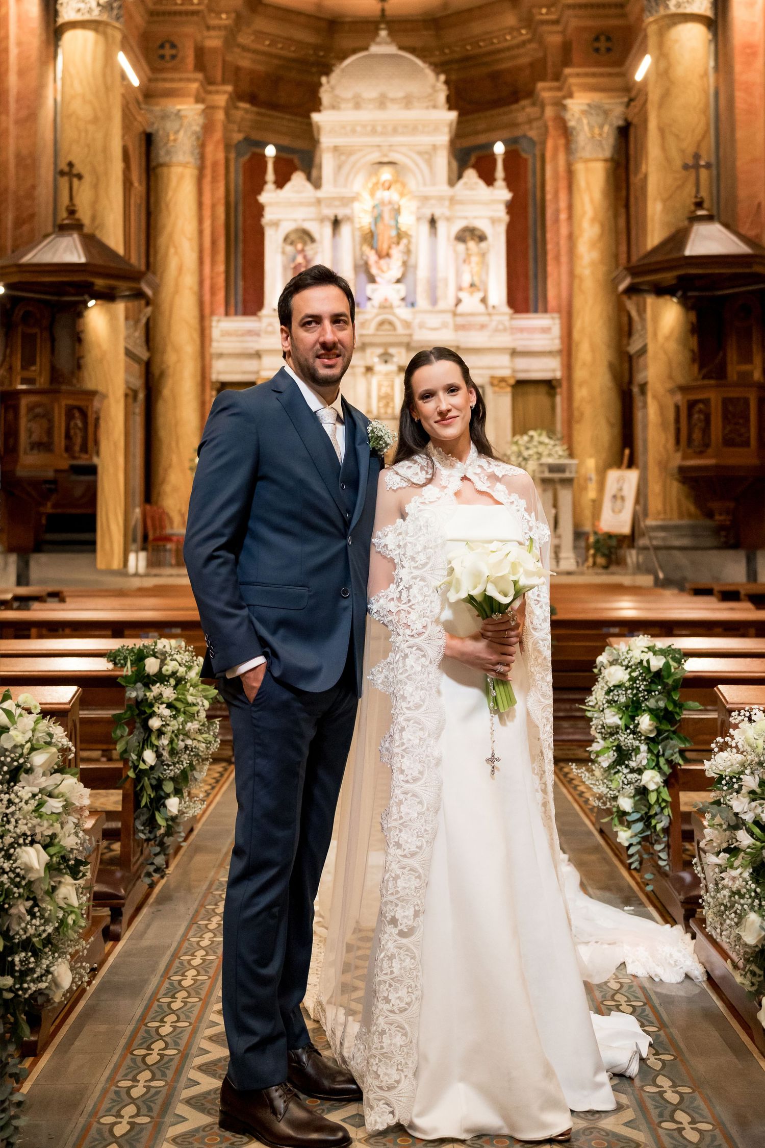 Bride and groom posing together at church altar, bride in lace gown with bouquet, groom in navy suit.