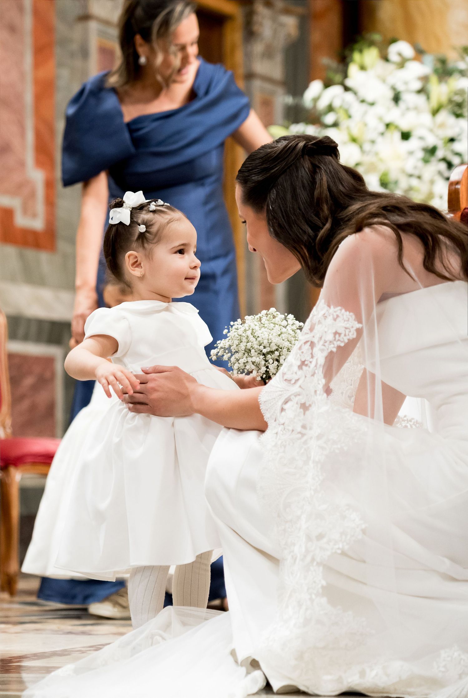 Bride in white gown crouches to interact with flower girl in white dress during elegant church wedding ceremony.