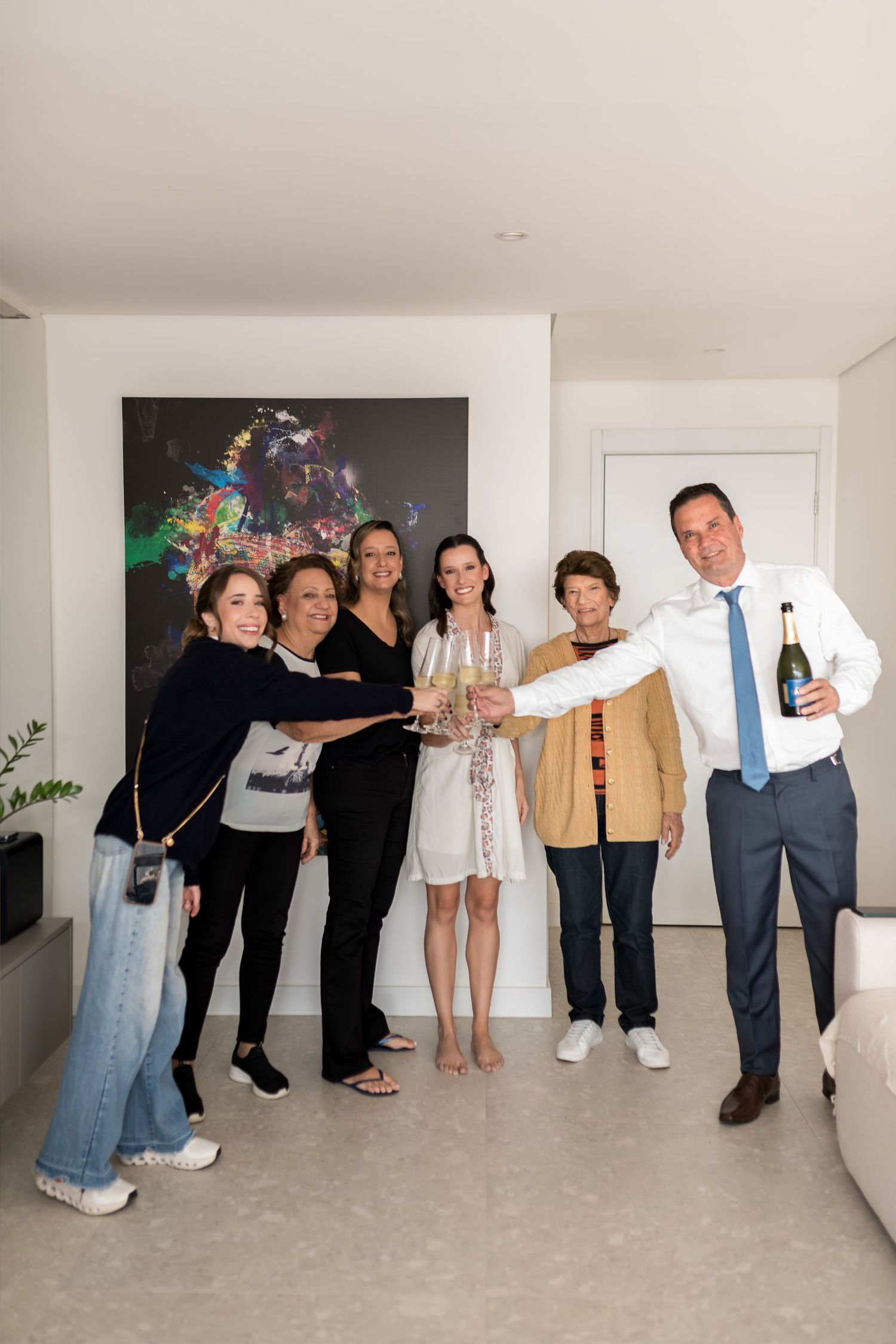 Group of six people toasting with champagne glasses and bottle in a modern living room with colorful artwork.