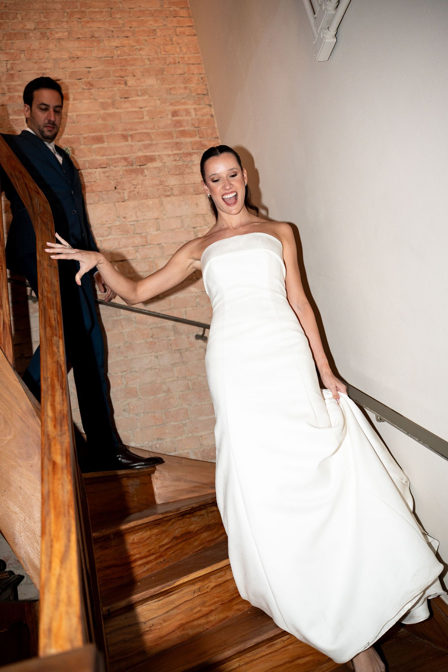 Bride in white strapless gown laughing on wooden staircase with groom in navy suit behind her.