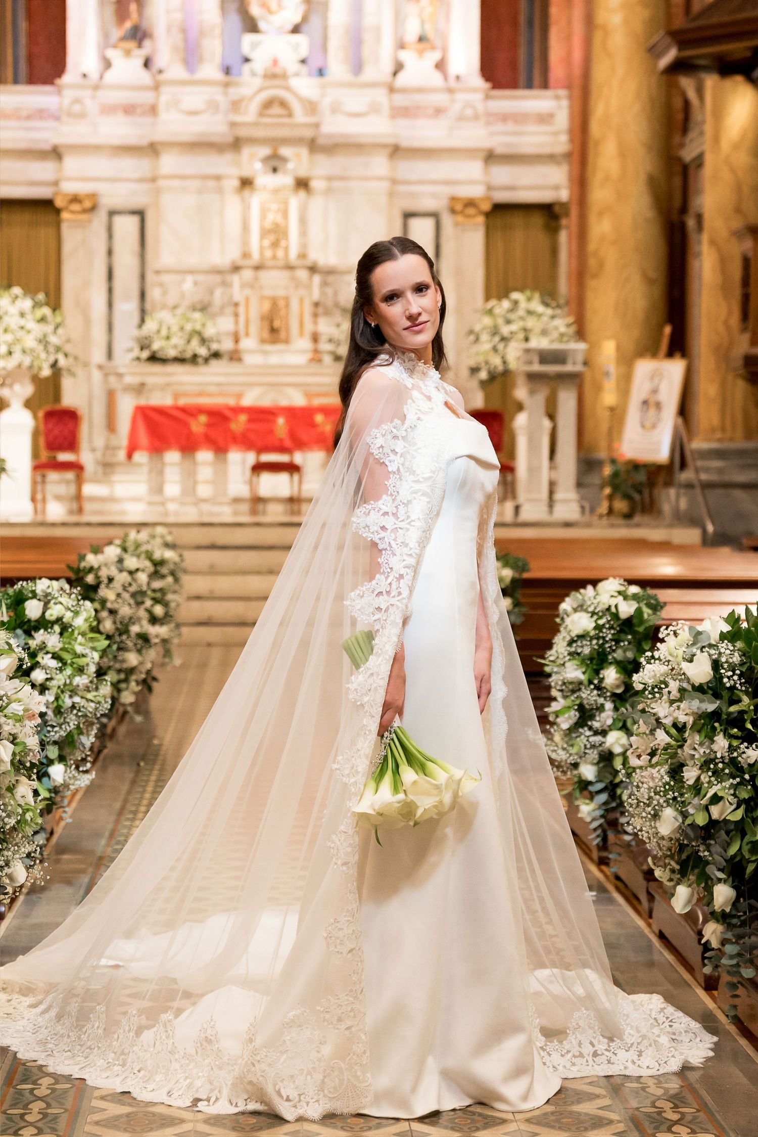 Bride in elegant white gown with lace cape veil holding tulip bouquet standing in ornate church aisle.