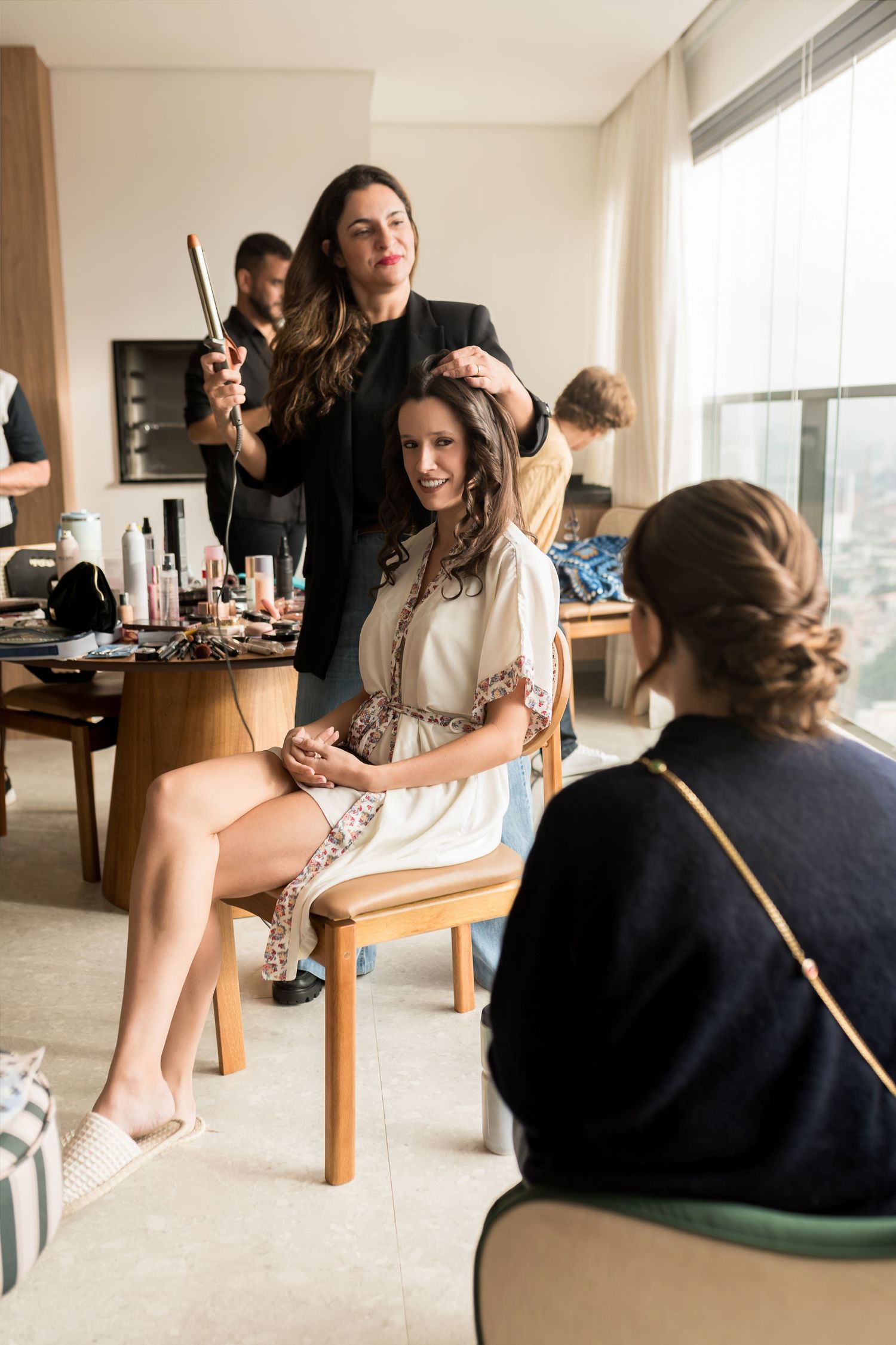 A hairstylist styling a seated woman's hair in a bright room with makeup products on a table nearby.