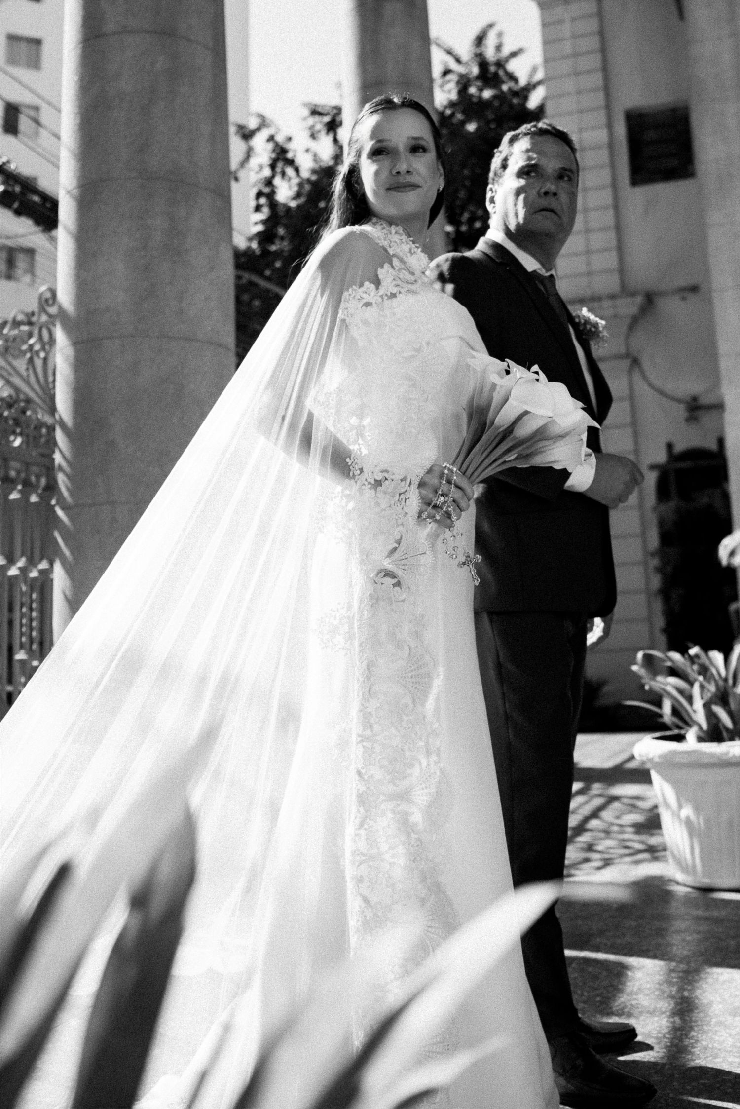 Black and white photo of a bride in lace gown and veil standing with a man in suit outside a church.