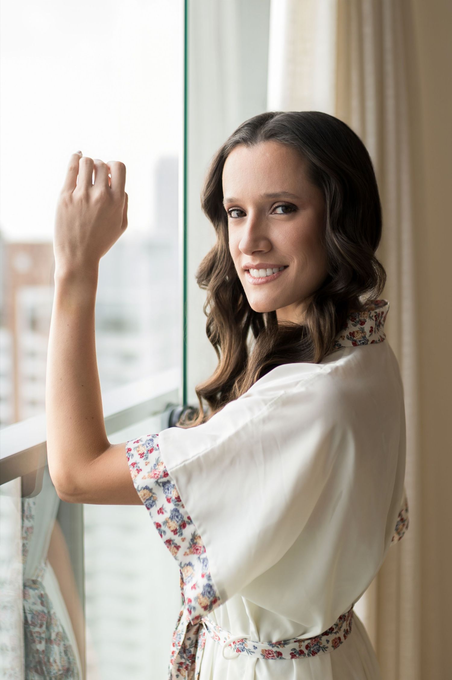 Smiling woman in floral robe standing by large window, looking over shoulder in bright modern setting.