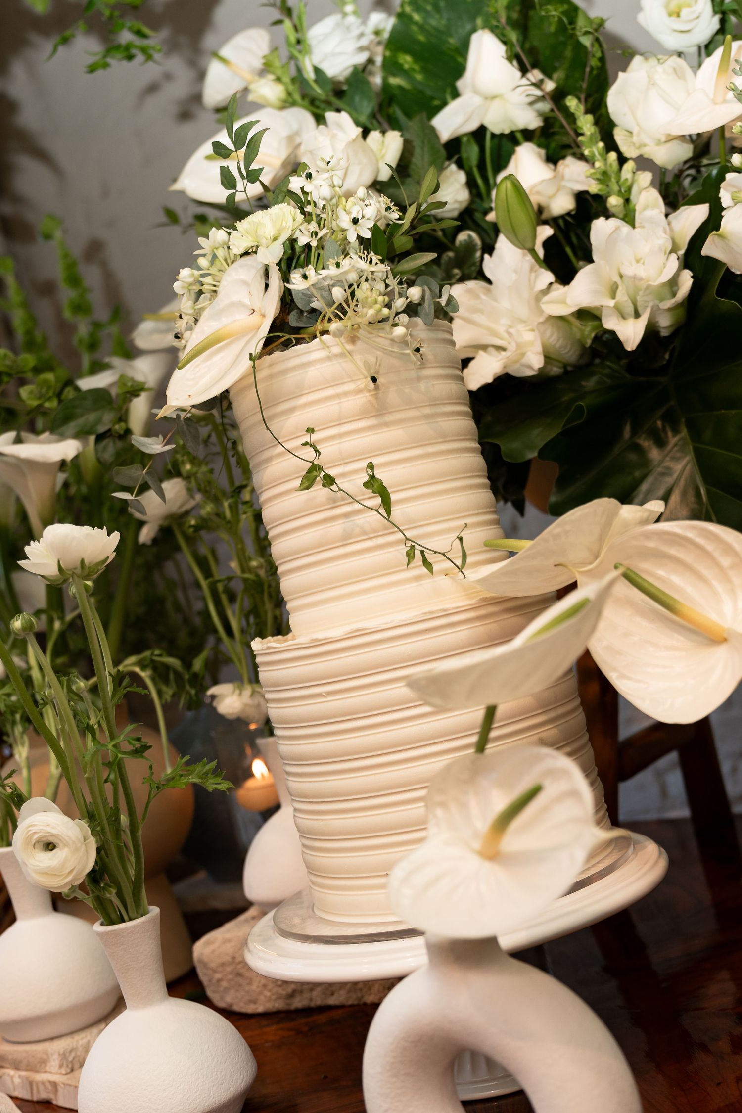 Elegant white tiered wedding cake adorned with floral decorations and surrounded by white flower arrangements on a wooden table.